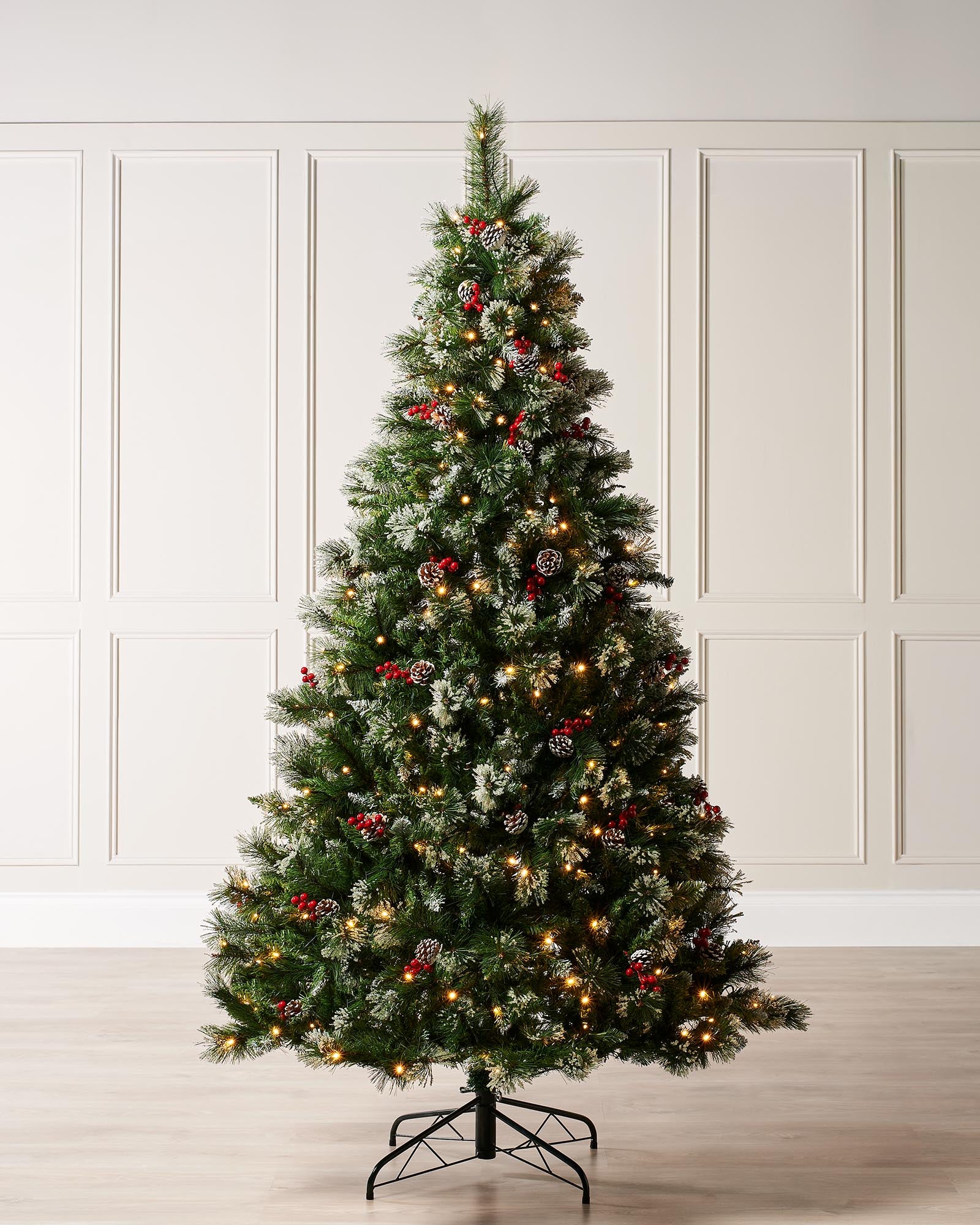 Pre-lit snow-frosted Christmas tree with warm white LED lights, decorated with frosted red berries and pinecones, standing on a metal stand against a plain background.