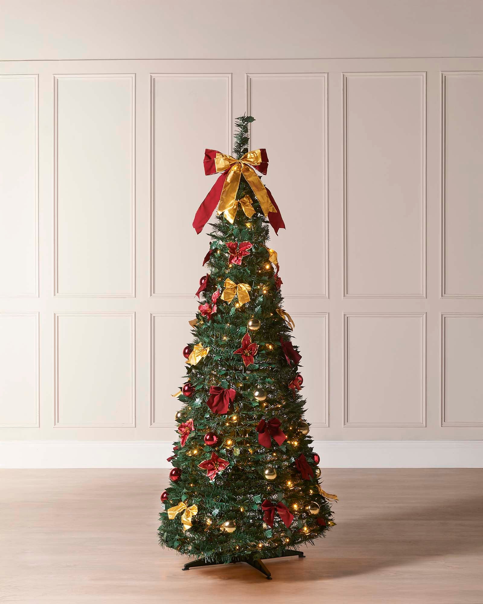 A 6-foot pre-lit pop-up Christmas tree decorated with red and gold baubles, ribbons, and flowers, featuring 150 warm white LED lights, displayed indoors against a white wall.