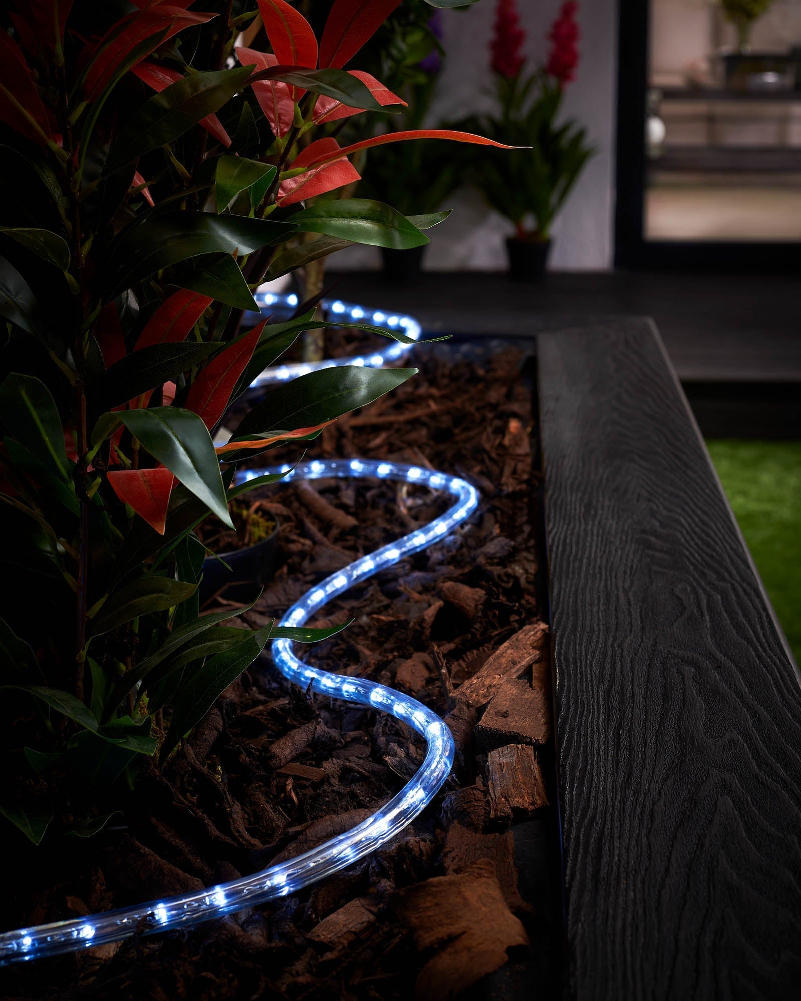 Connectable white LED rope light installed around a garden bed, displaying bright light in a dark environment.