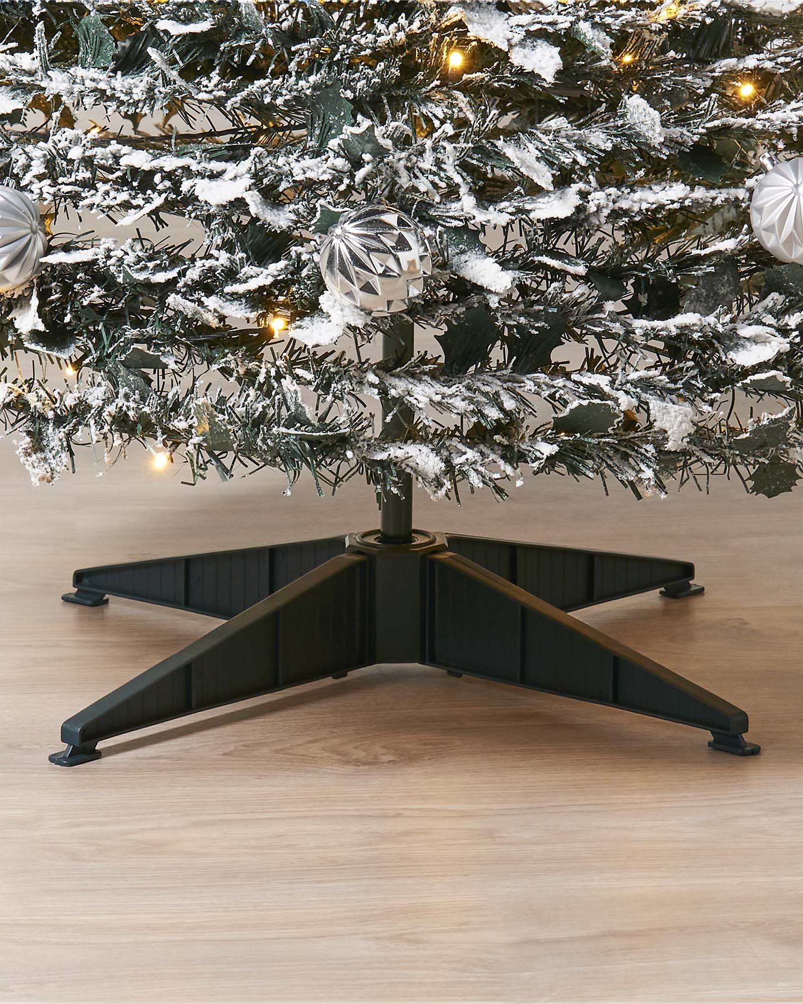 Black adjustable Christmas tree stand with a decorated tree on a wooden floor. 6 ft pre-lit decorated pop-up snow flocked Christmas tree with 150 warm white LEDs, white and silver baubles, and a snow-flocked finish—easy setup, creating a cozy indoor winter wonderland centerpiece.