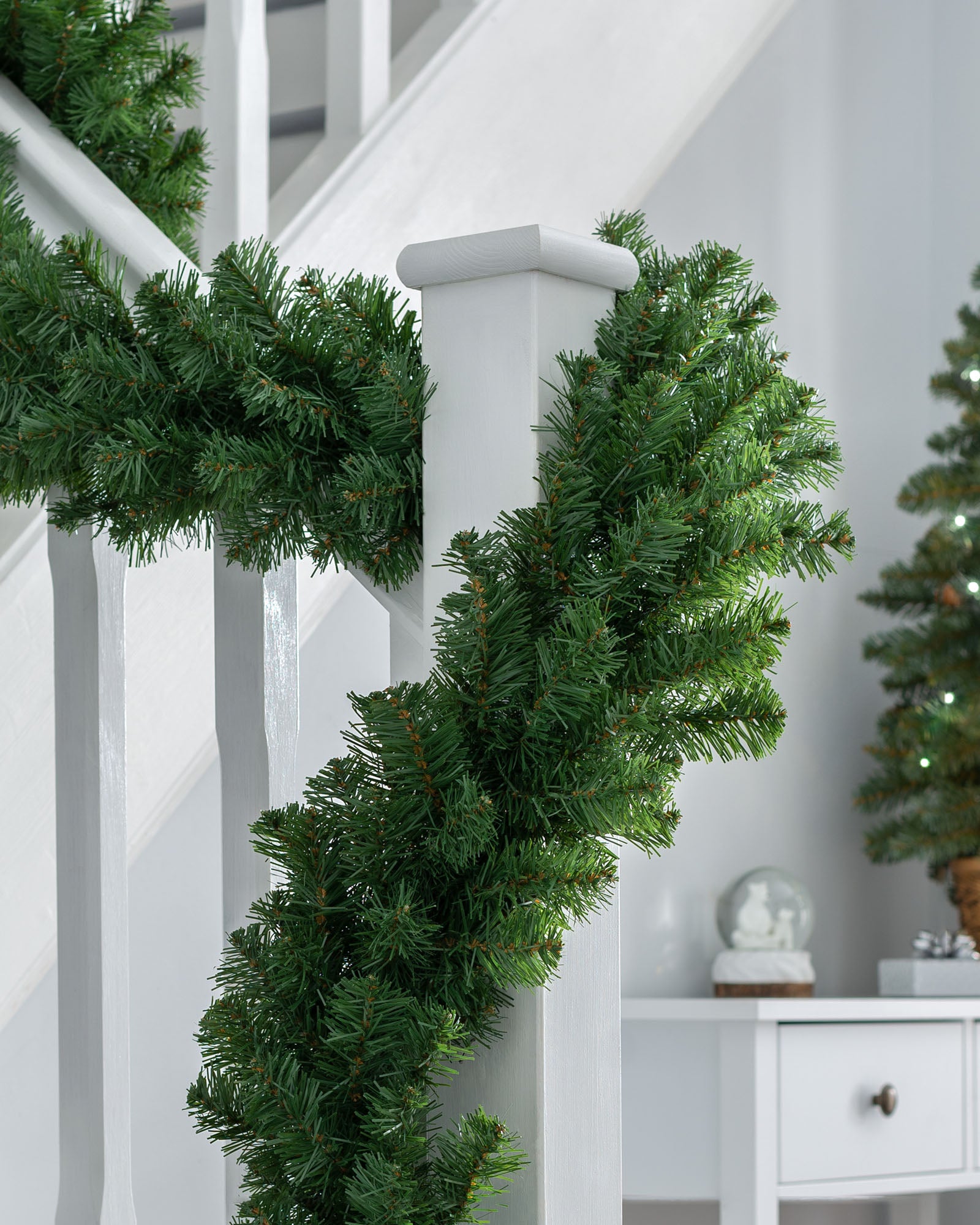 A green, extra thick pine garland draped around a staircase banister, with a white railing and a decorated Christmas tree in the background.