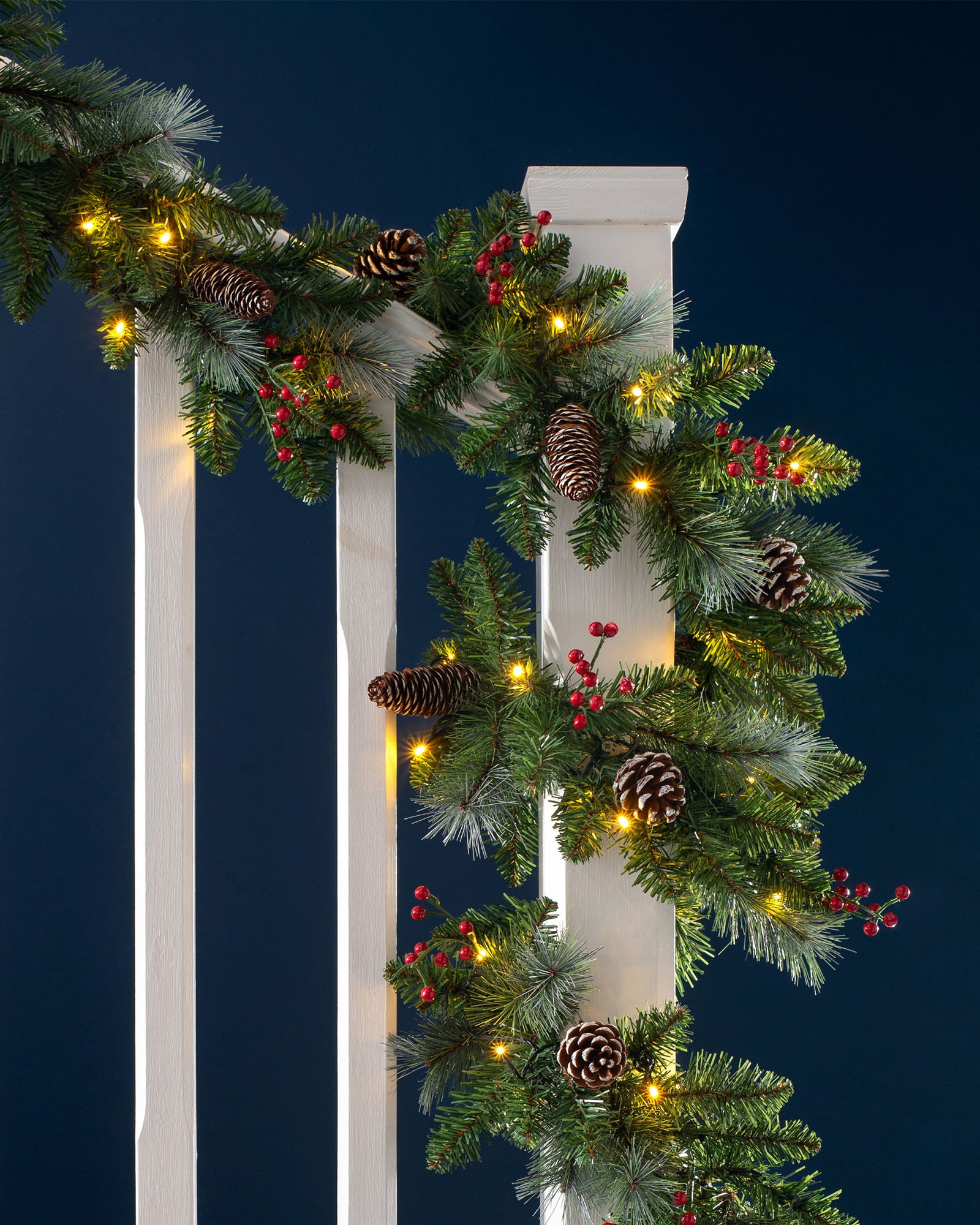 A festive pre-lit garland with frosted pinecones and red berries, illuminated by warm white LED lights, draped across a stair railing.