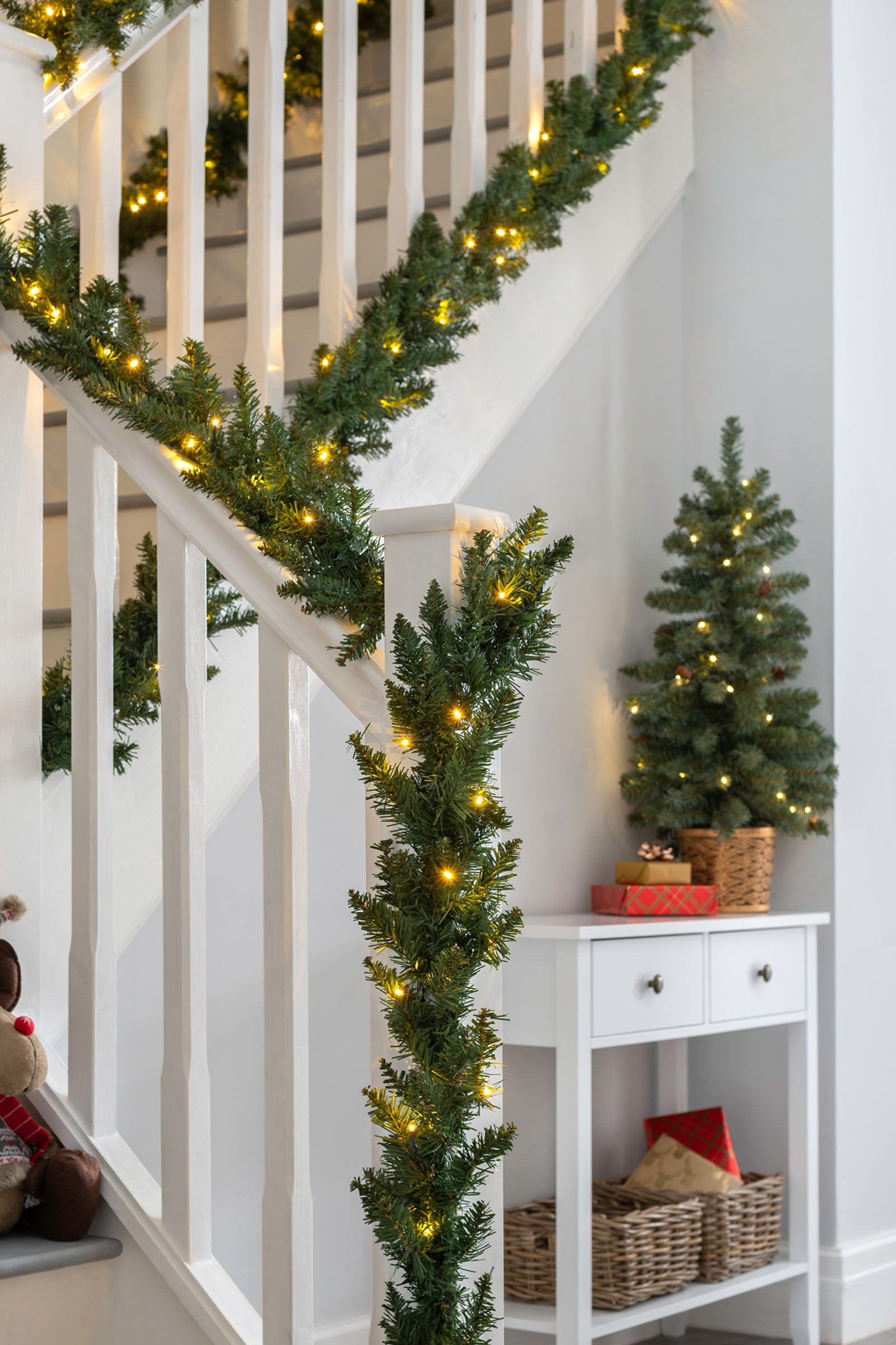A green garland with warm white LED lights draped over a staircase with a white railing, leading up to a brighter area.