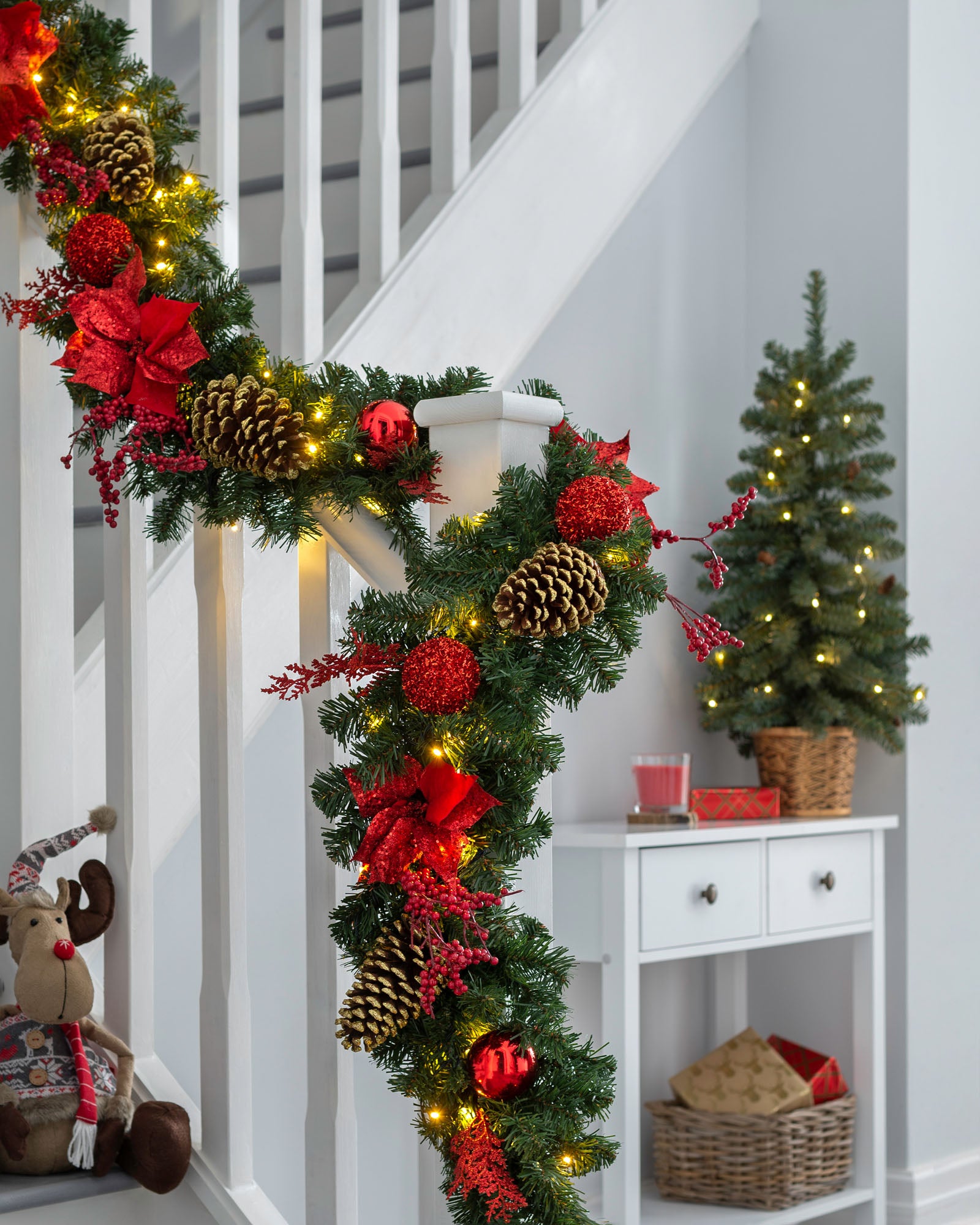 A festive pre-lit red decorated extra thick garland with warm white LED lights, pine cones, and red ornaments draped across a banister.