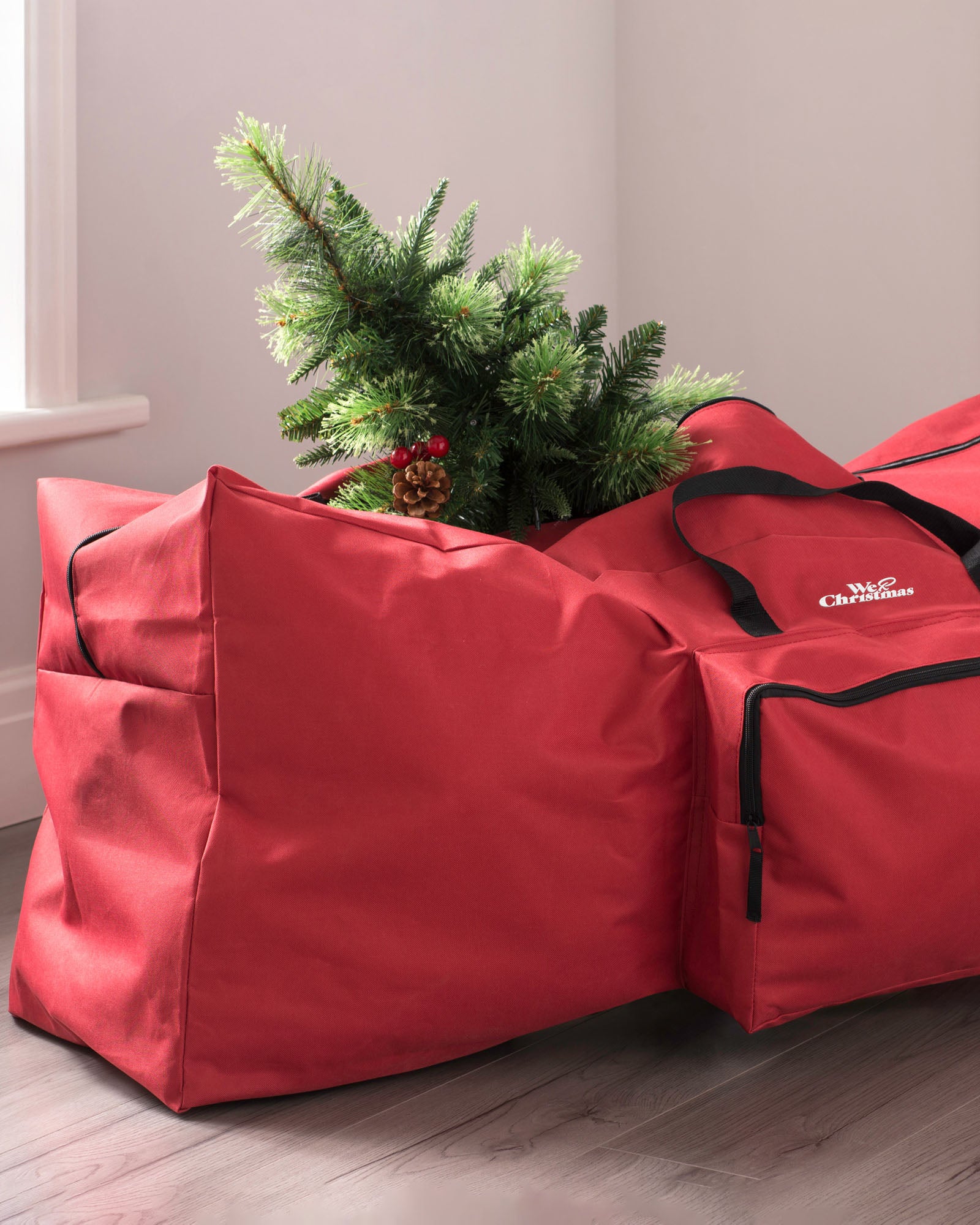 A red Christmas tree storage bag with a Christmas tree being stored inside it, placed on a wooden floor against a white wall.