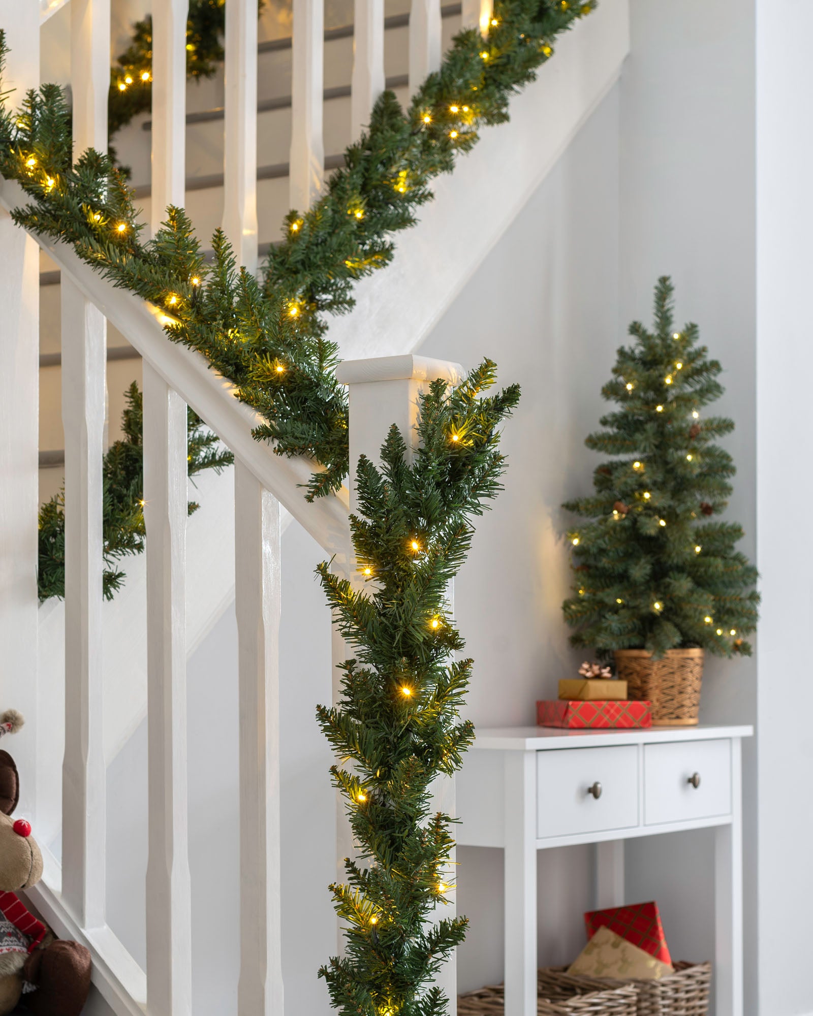 A green garland with warm white LED lights draped over a staircase, with a white wall and decorative accents in the background.
