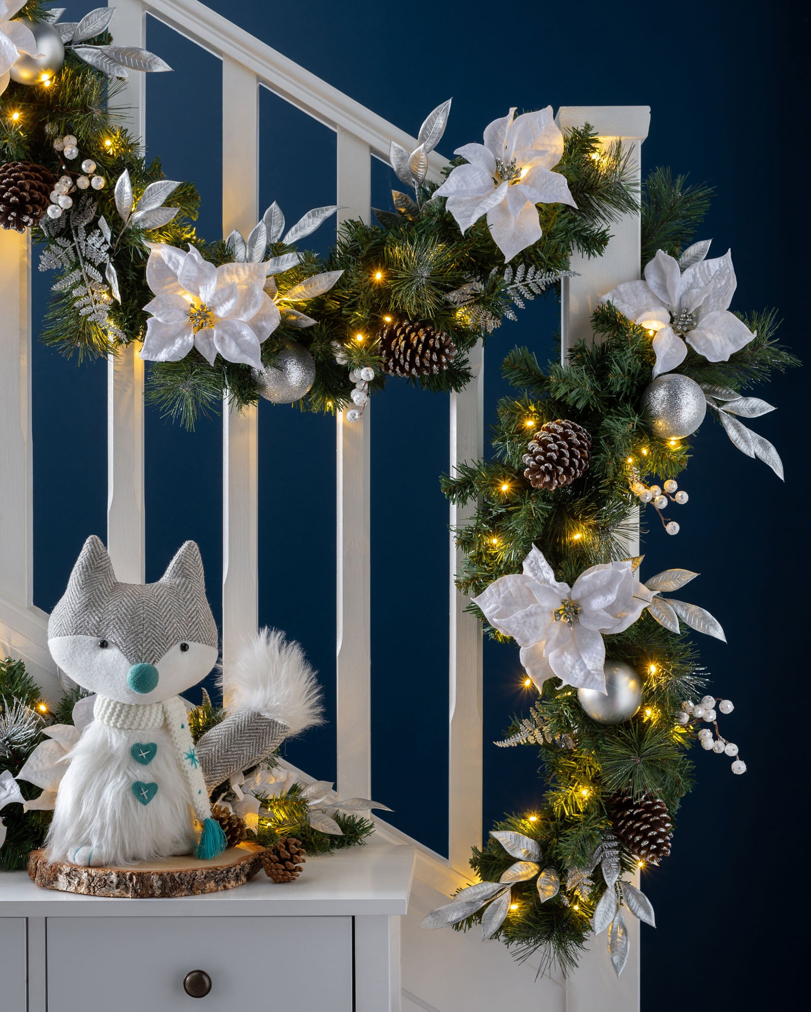 A decorated garland with silver and white elements, including LED lights, pinecones, and faux flowers, displayed on a dark background.