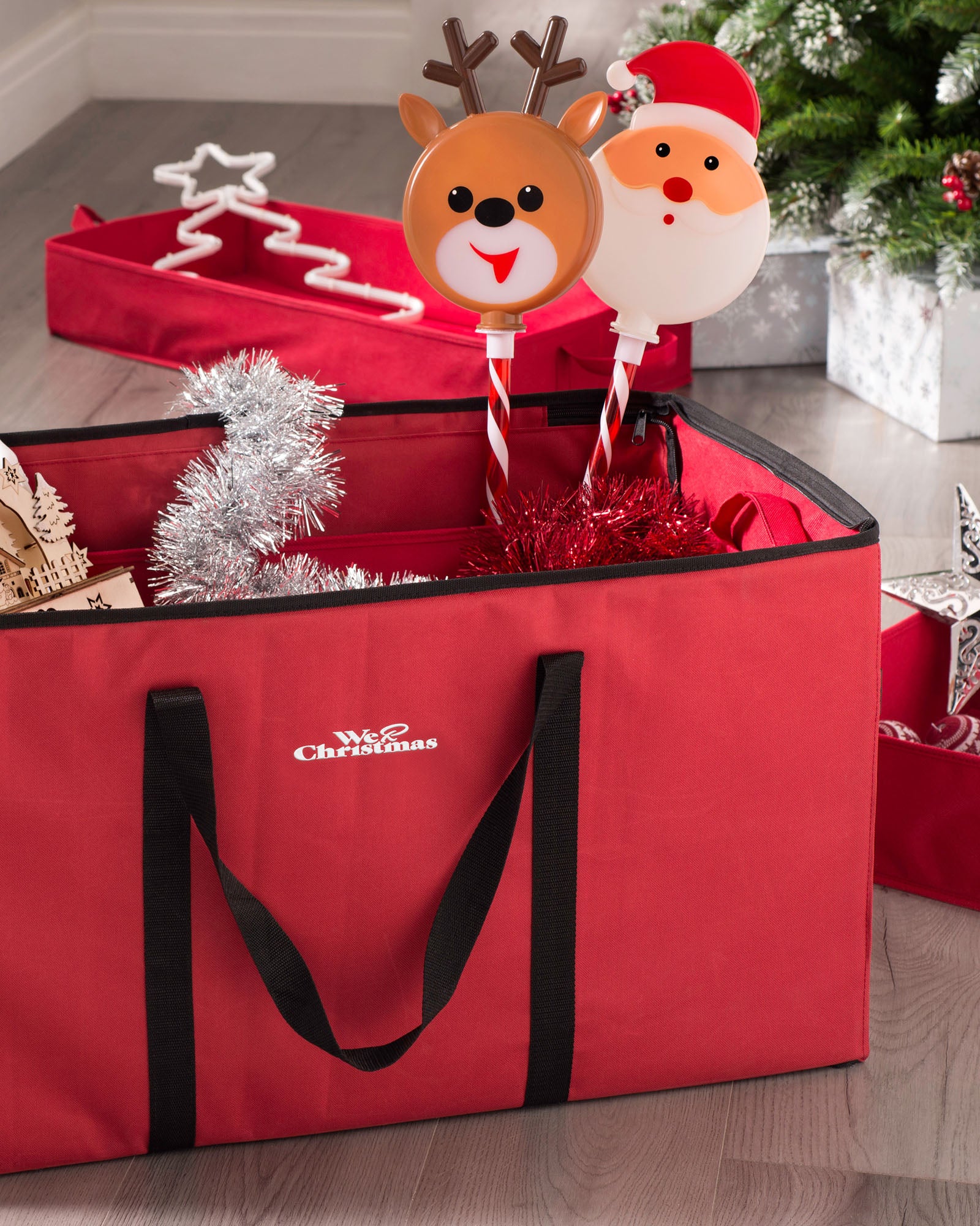 A red fabric ornament and decorations storage box with a festive design, containing various Christmas decorations such as a reindeer and Santa figurine, tinsel, and other trinkets.