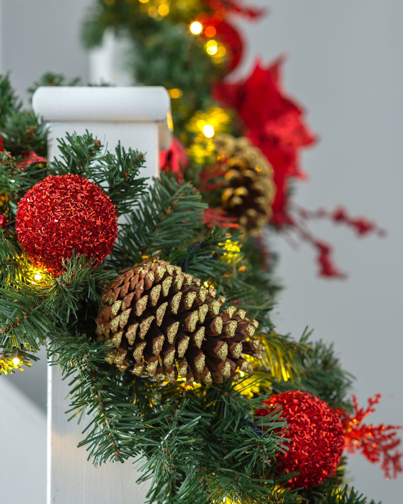 A festive pre-lit red decorated extra thick garland with warm white LED lights, pine cones, and red ornaments draped across a banister.