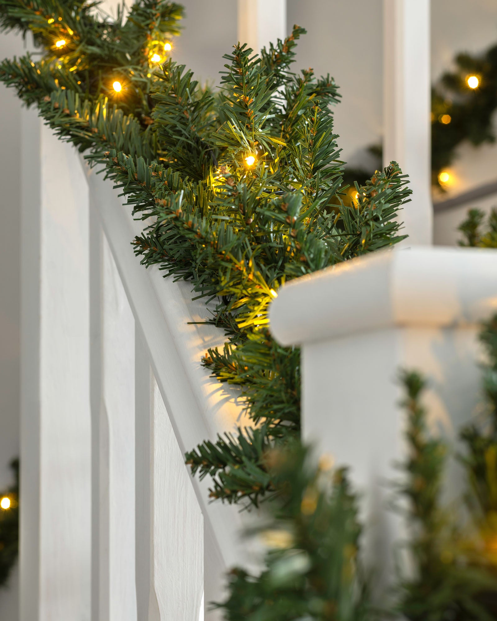 A green garland with warm white LED lights draped over a staircase, with a white wall and decorative accents in the background.