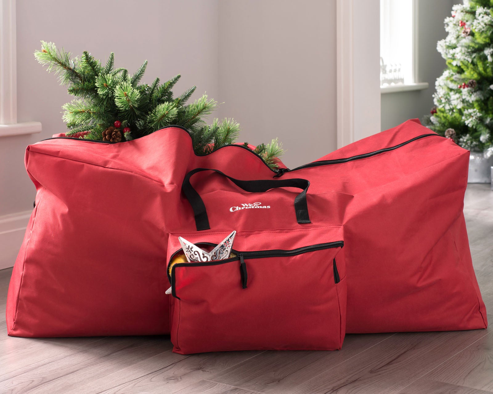 A red Christmas tree storage bag with a Christmas tree being stored inside it, placed on a wooden floor against a white wall.