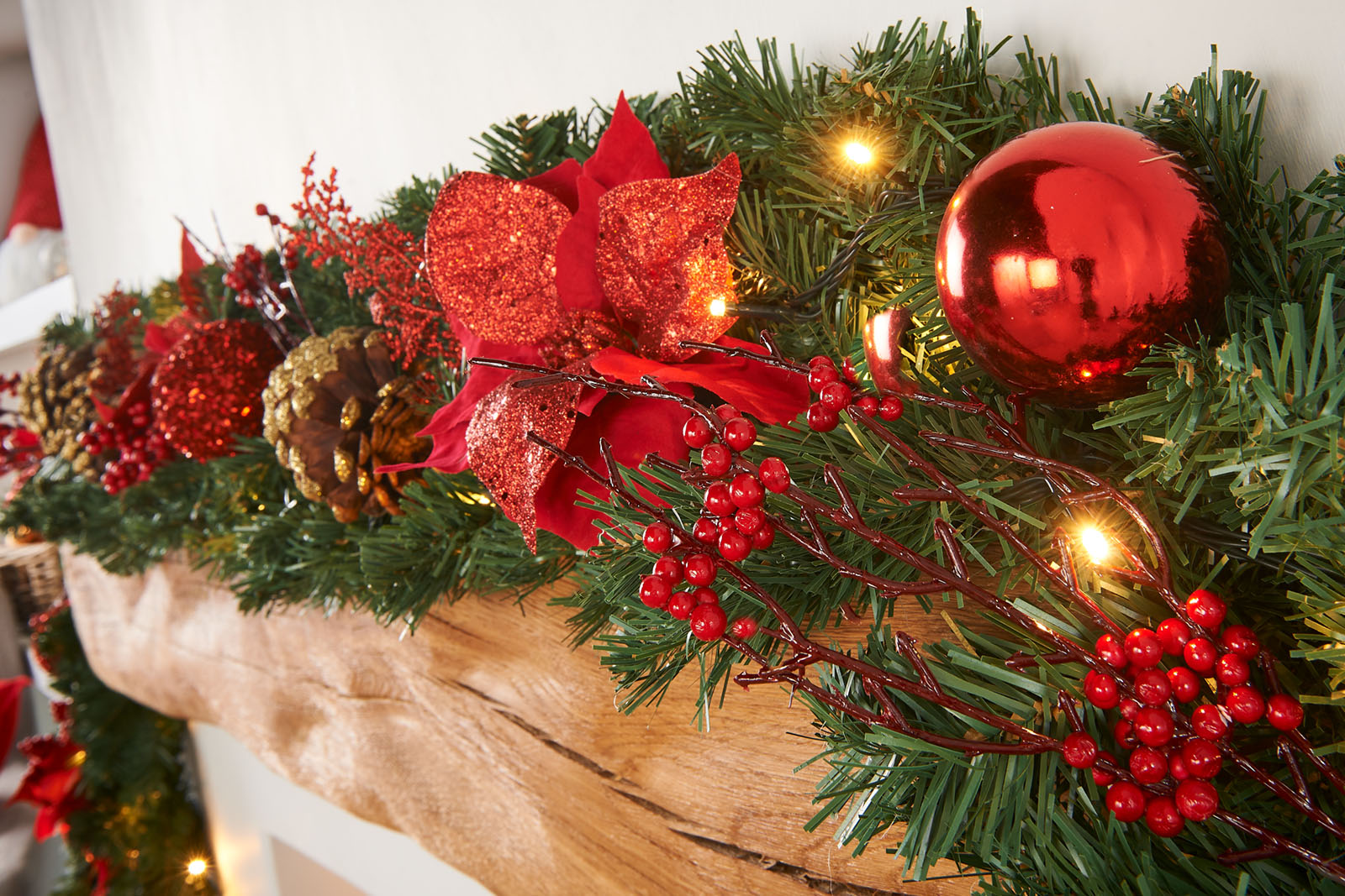 A close-up of a pre-lit extra thick decorated Christmas garland with red and gold elements, including berries, pine cones, baubles, and glittery decorations, illuminated by warm white LED lights.