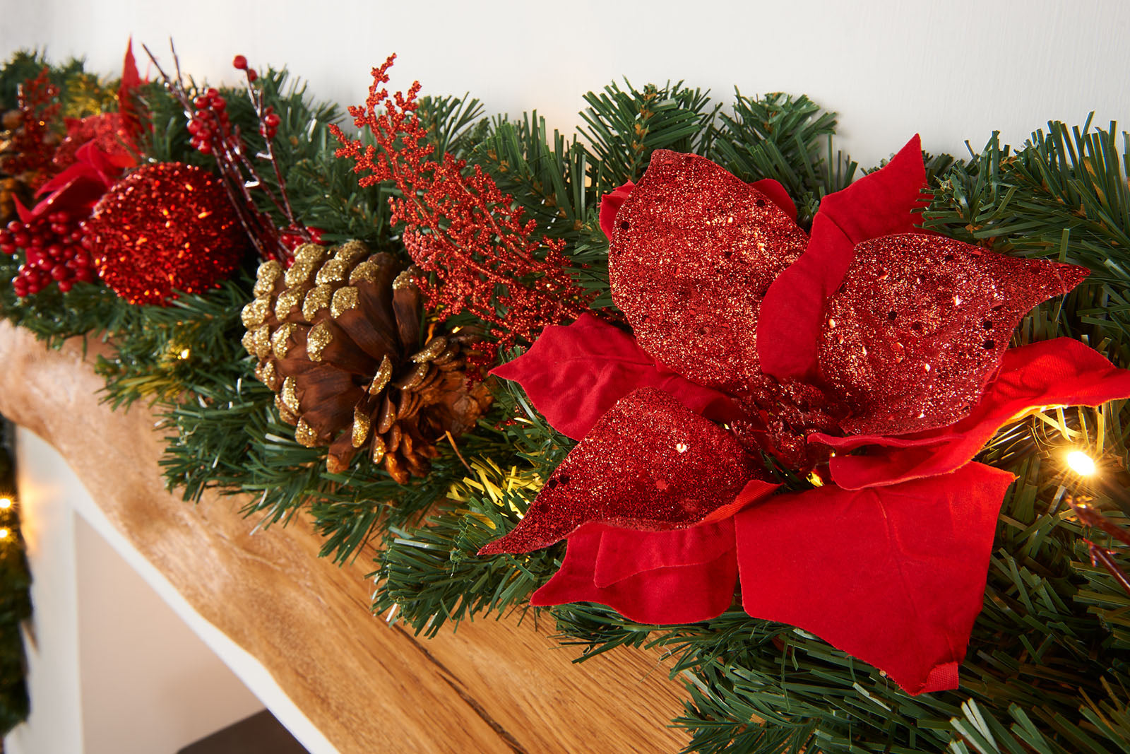 A close-up of a pre-lit extra thick decorated Christmas garland with red and gold elements, including berries, pine cones, baubles, and glittery decorations, illuminated by warm white LED lights.