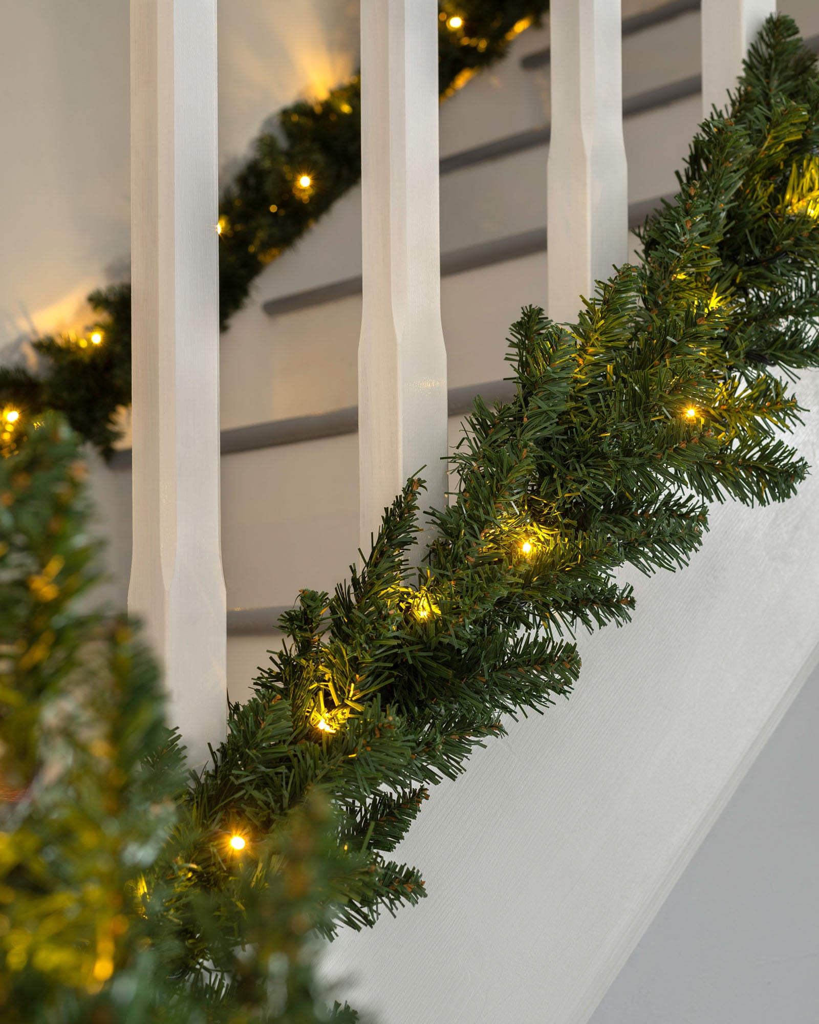 A green garland with warm white LED lights draped over a staircase, with a white wall and decorative accents in the background.