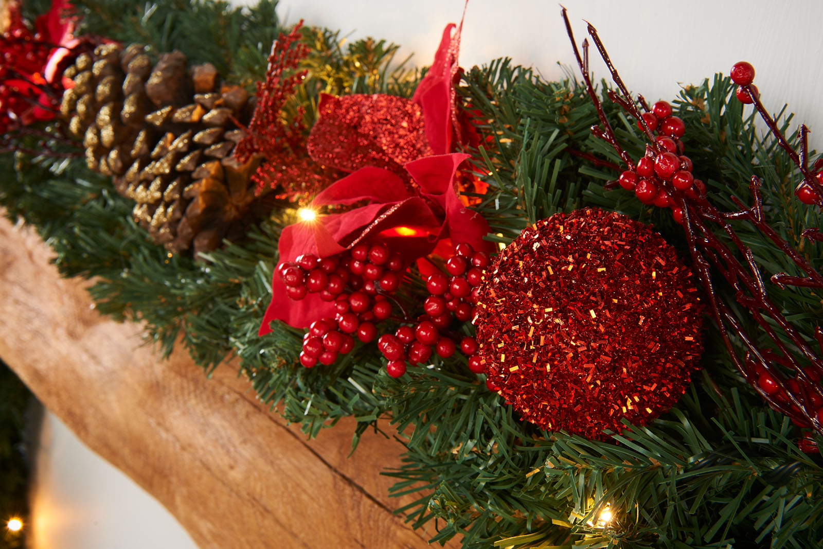 A close-up of a pre-lit extra thick decorated Christmas garland with red and gold elements, including berries, pine cones, baubles, and glittery decorations, illuminated by warm white LED lights.