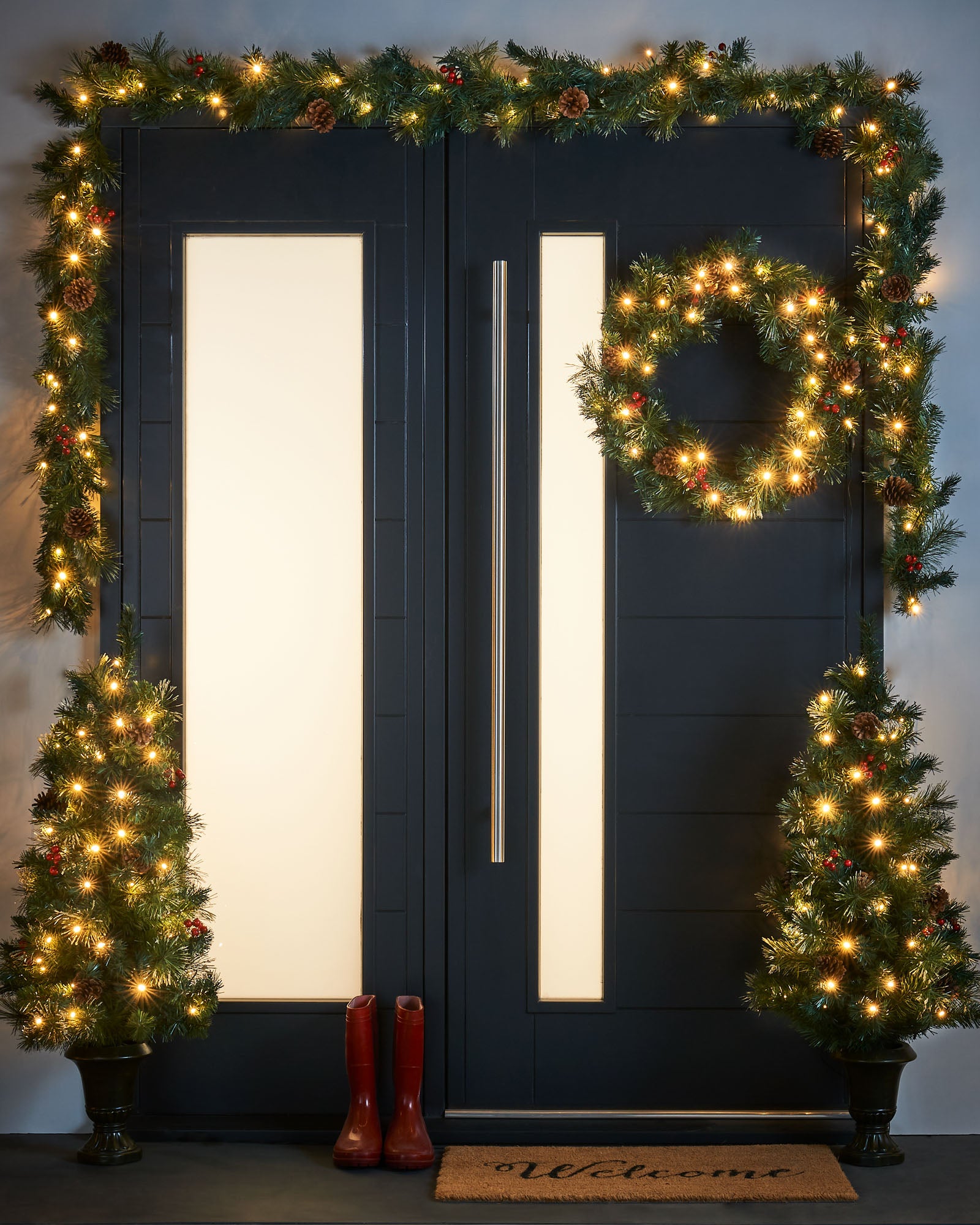 Pre-lit decorative Christmas trees, wreath, and garland arranged on a dark doorway, illuminated with warm white LED lights. Pre-lit Christmas decoration set including two potted trees, a wreath, and a garland—all featuring warm white LED lights, red cluster berries, pinecones, and frosted PVC tips—ideal for creating a coordinated festive display at a home entrance.