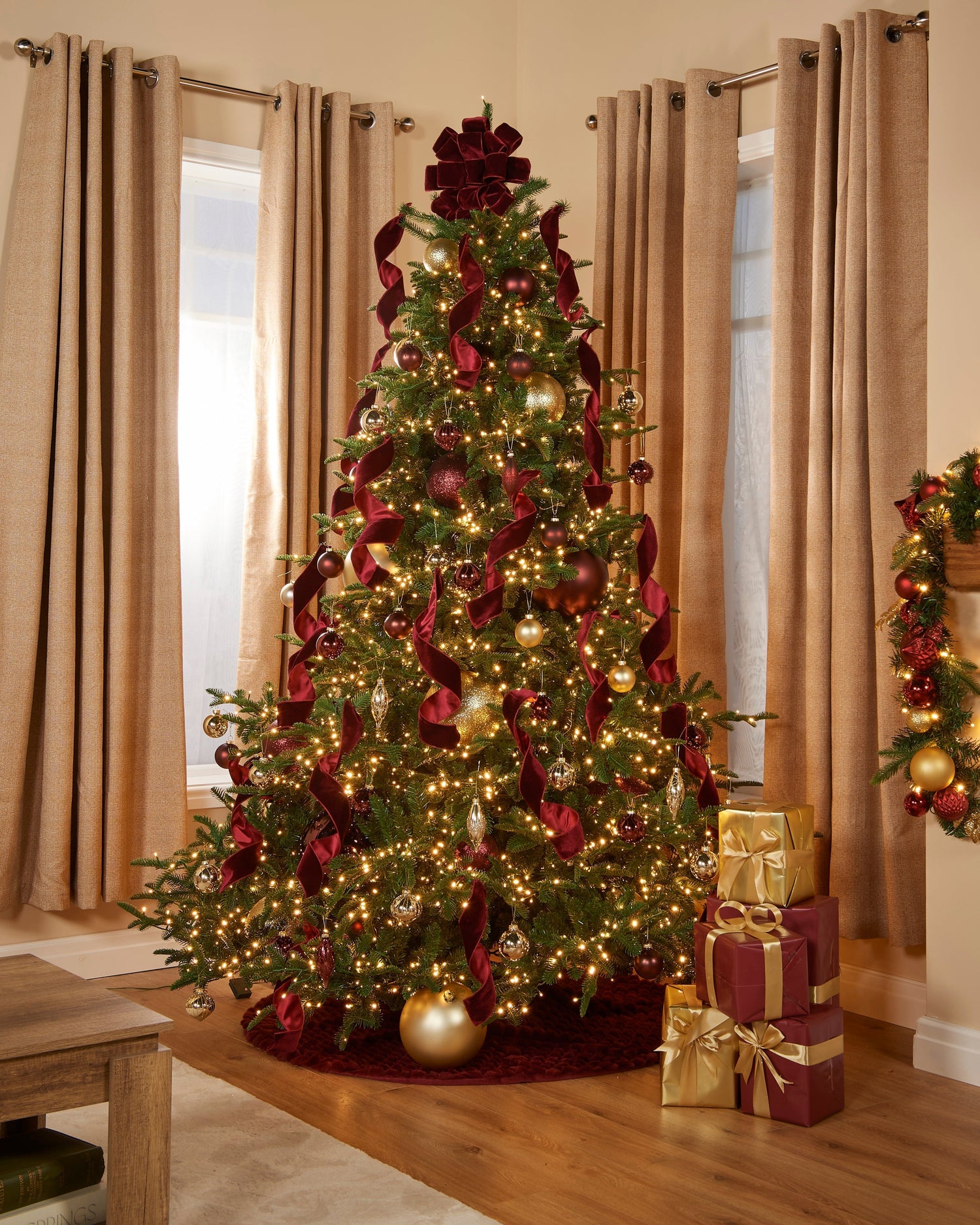 A luxury, pre-lit Christmas tree in a studio adorned with burgundy and gold toned ornaments and decorations.
