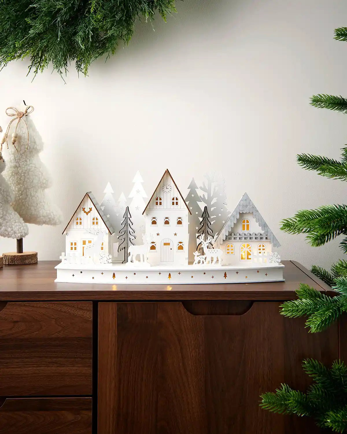 Scenic full image of 40cm white wooden village with warm white lighting on dark surface in front of white wall