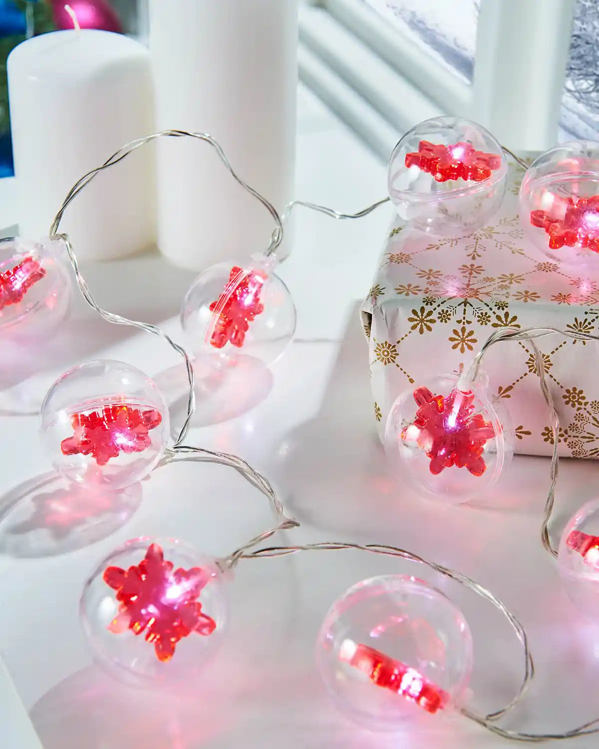 Red snowflake bauble LED string lights styled on a white table beside candles and wrapped gifts