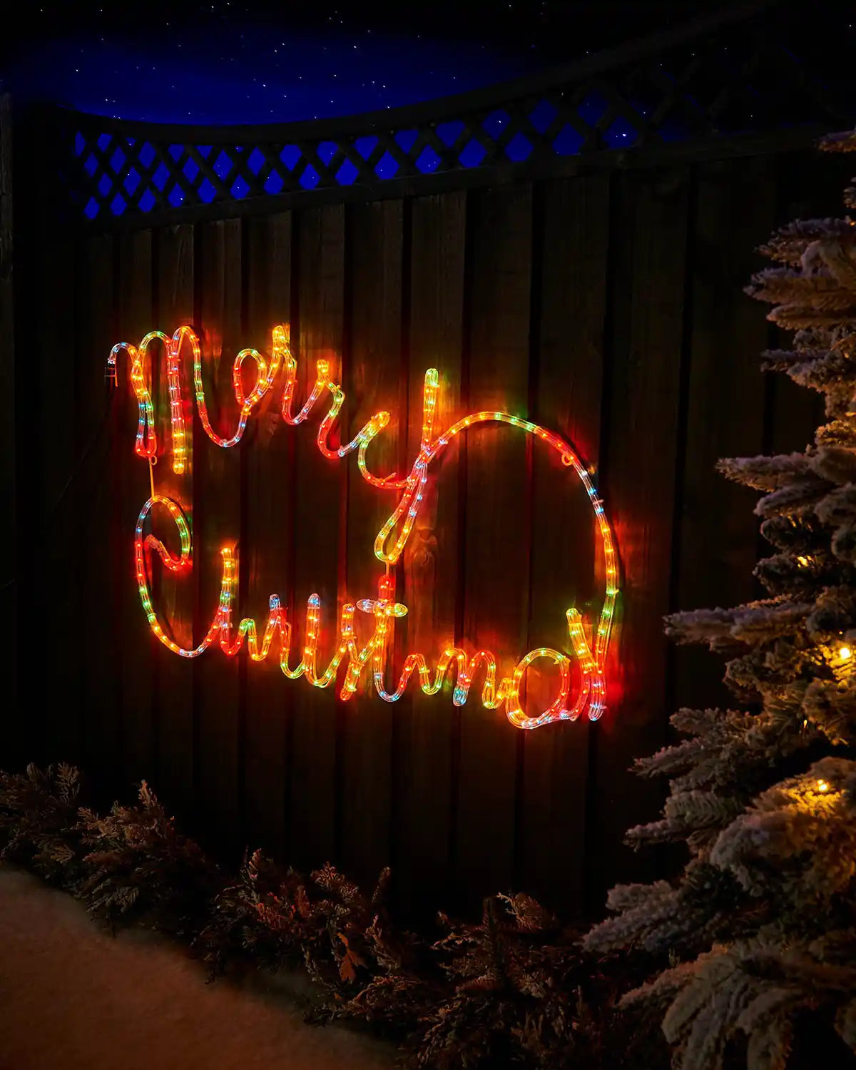 Multi-coloured Merry Christmas rope light display on a garden fence at night