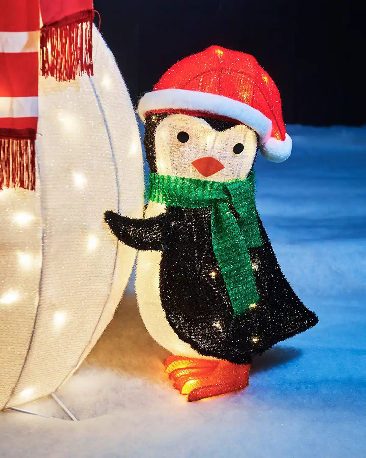 Close-up of a pre-lit penguin wearing a red Santa hat and green scarf beside the snowman.