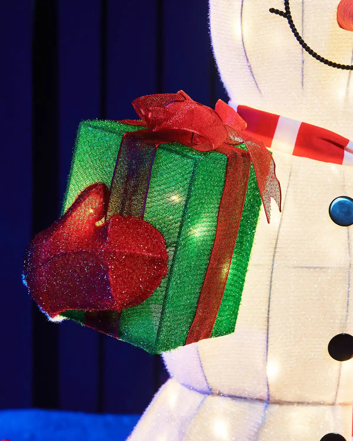 Close-up of the snowman holding a green gift with red ribbon and warm white lights.
