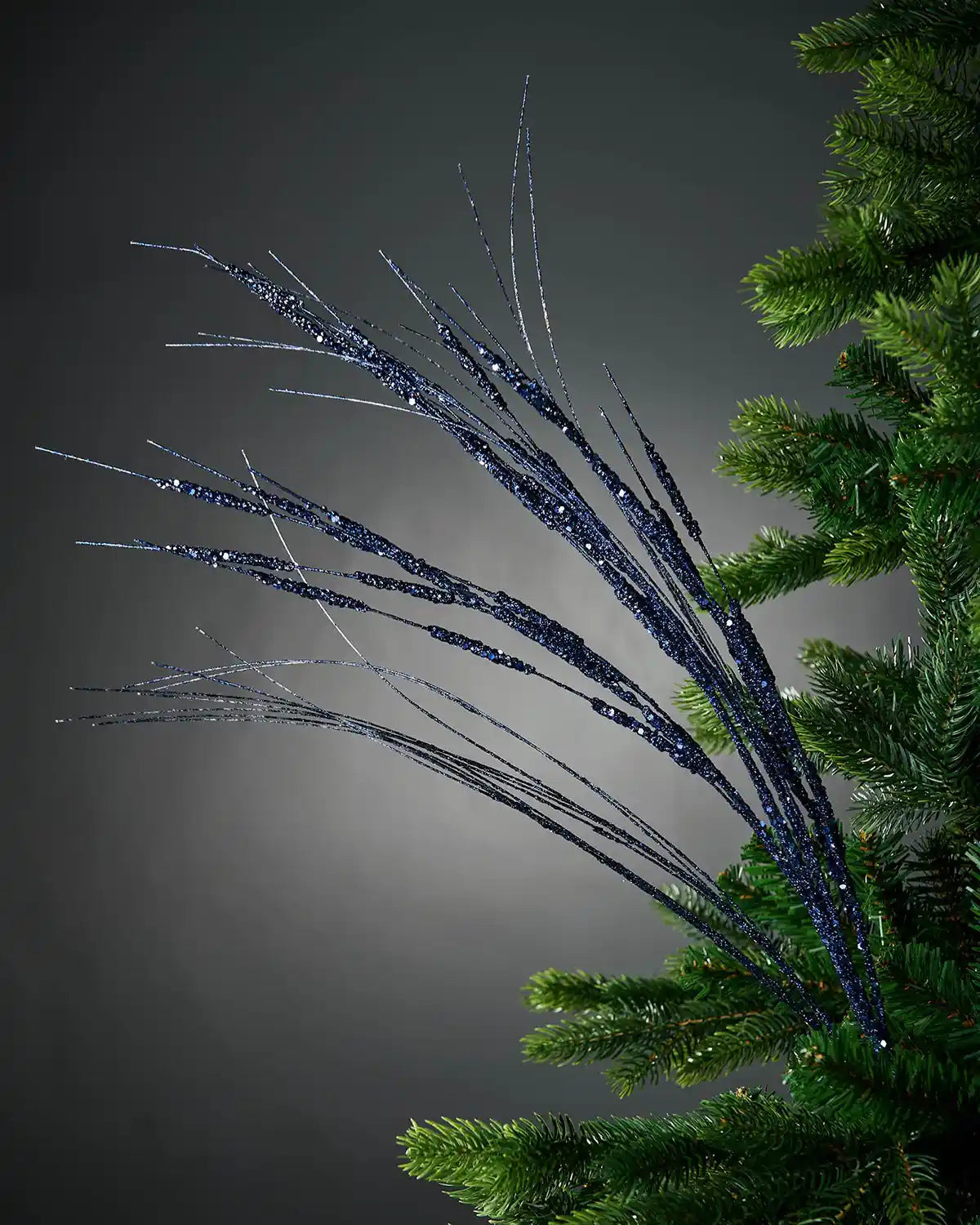 Full image of 100cm navy blue glitter wispy tree spray attached to Christmas tree in front of grey wall background
