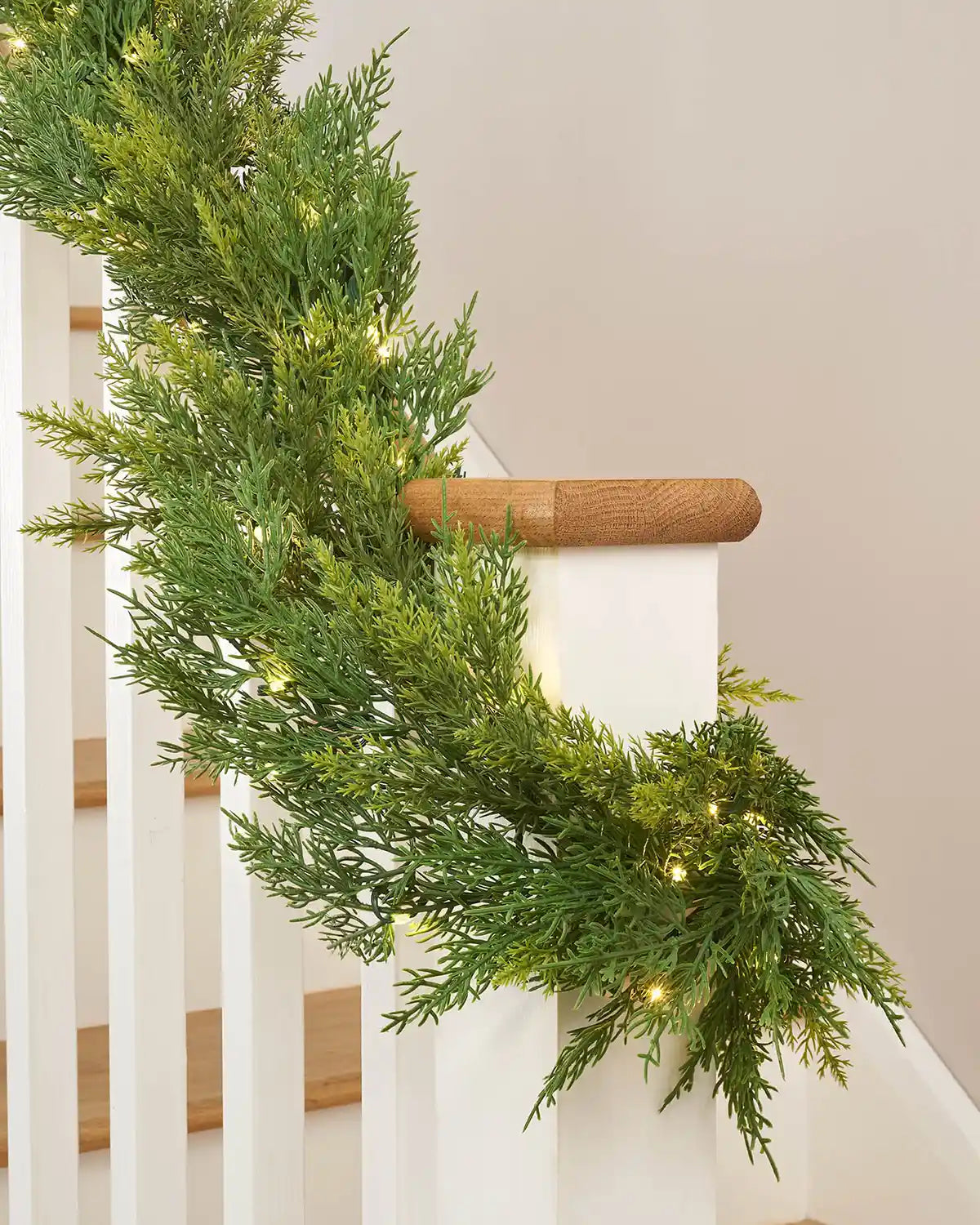 Close-up of 12ft pre-lit cypress fir garland with warm white light setting wrapped around white wooden staircase banister in front of white wall