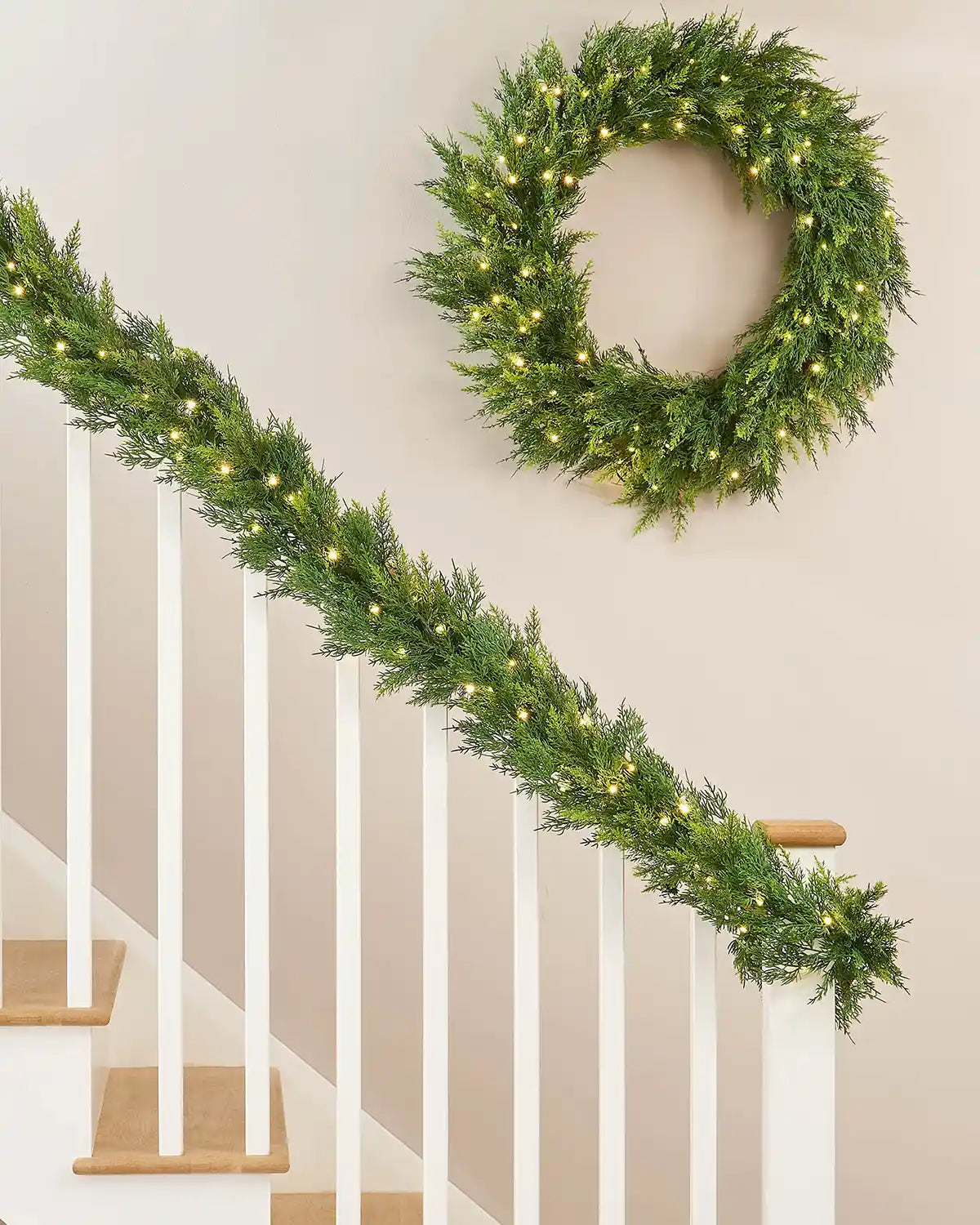 12ft pre-lit cypress fir garland placed on white wooden staircase banister with warm white light setting and matching wreath above on white wall
