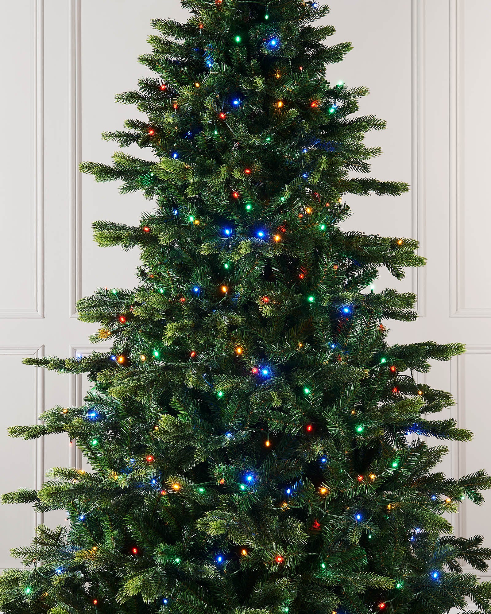 Tall Christmas tree with colorful lights against a white paneled background.