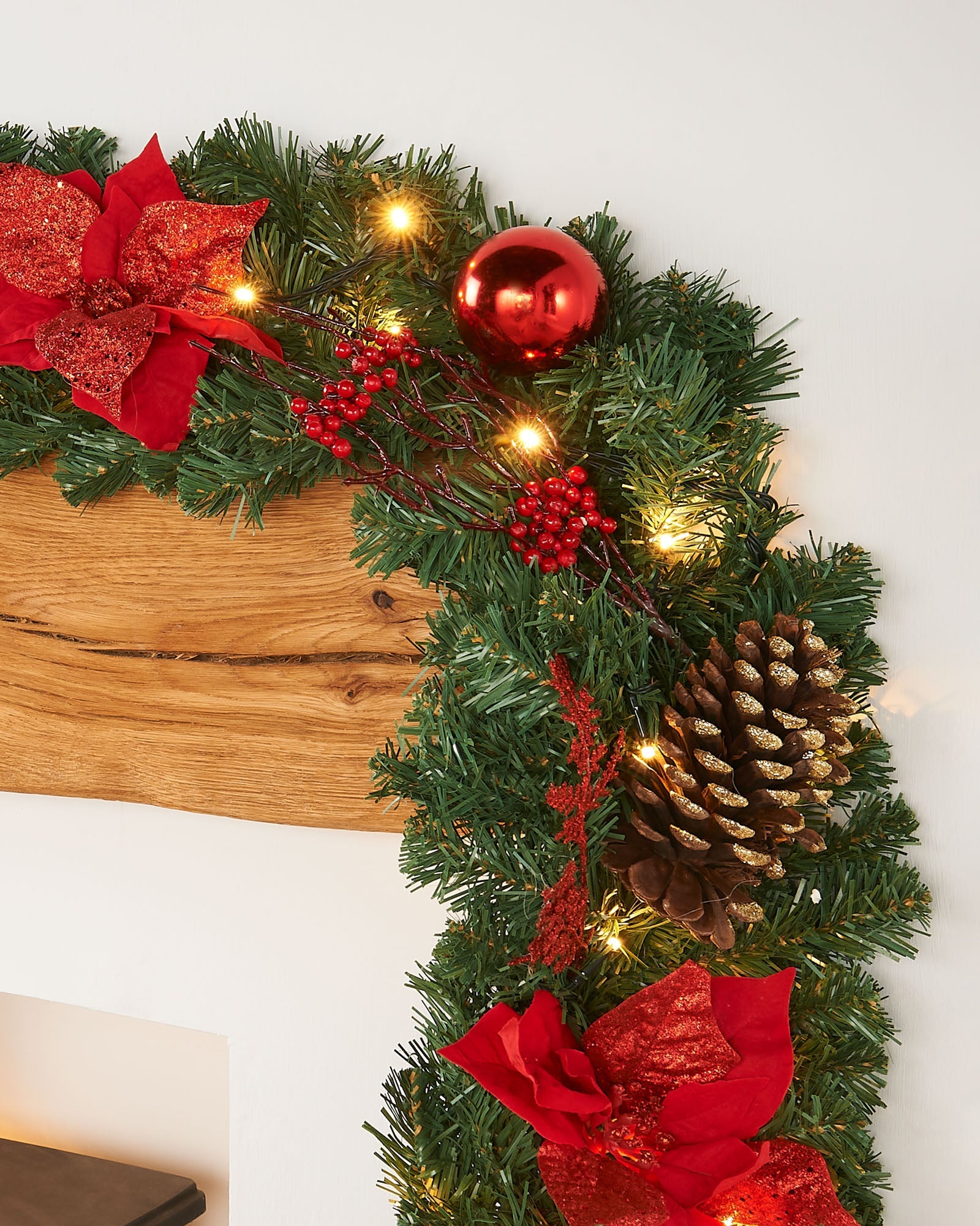 A close-up of a pre-lit extra thick decorated Christmas garland with red and gold elements, including berries, pine cones, baubles, and glittery decorations, illuminated by warm white LED lights.