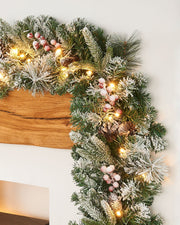 A close-up of a pre-lit, extra thick, mixed pine snow flocked garland with pinecones and berries, featuring warm white LED lights.