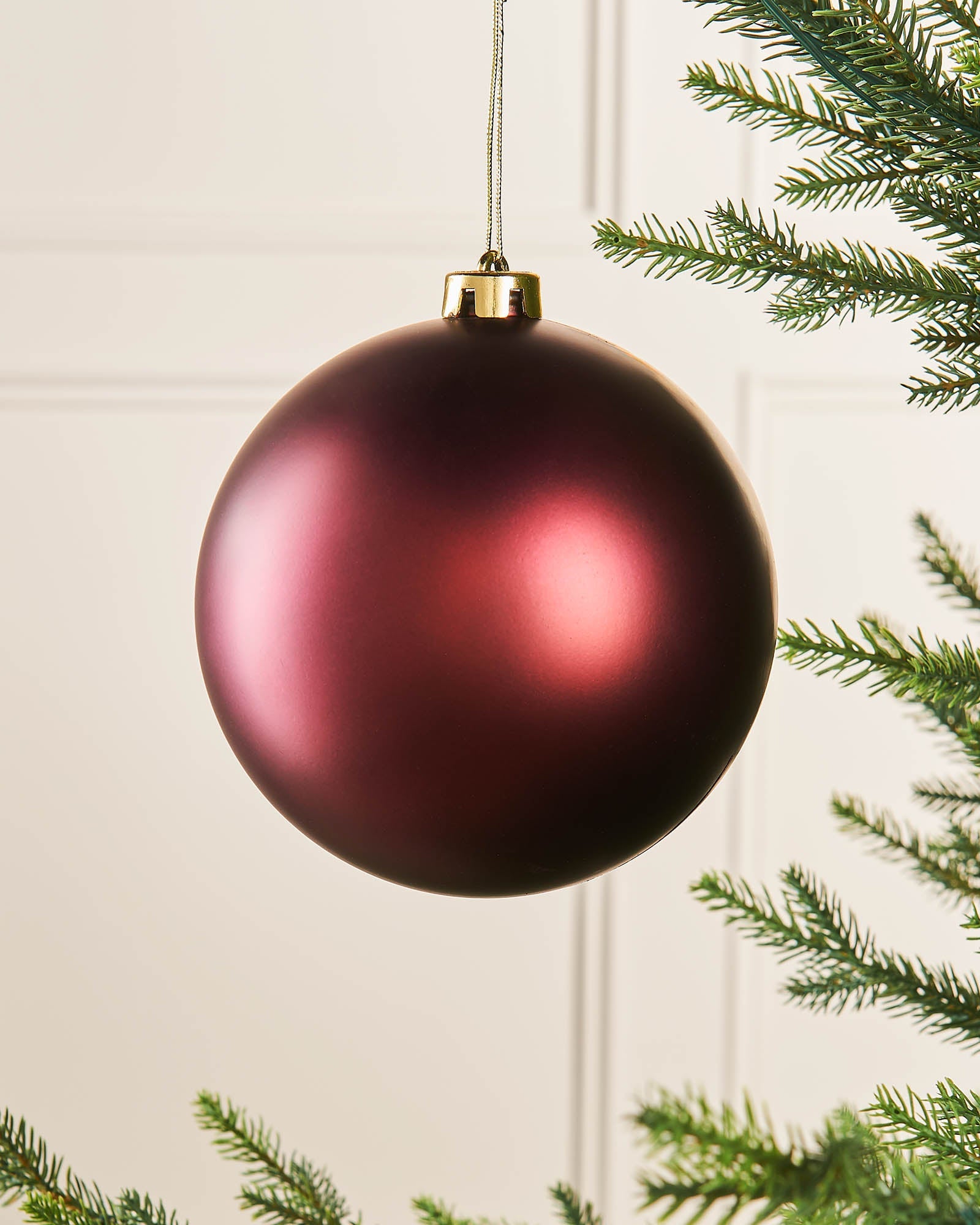 A burgundy oversized matte shatterproof bauble hanging from a string against a white background with green foliage at the sides.