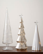Three decorative Christmas trees made of glass, metal, and ceramic on a wooden surface with a neutral background.