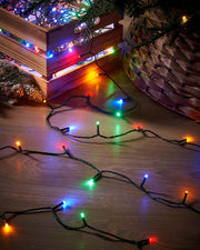 Multicolored string lights on a wooden surface with a Christmas tree in the background