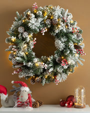 A pre-lit, extra thick, mixed pine snow flocked wreath with pinecones and berries, displayed against a warm background, with Christmas decorations and a toy figure to the side.