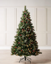 Pre-lit snow-frosted Christmas tree with warm white LED lights, decorated with frosted red berries and pinecones, standing on a metal stand against a plain background.