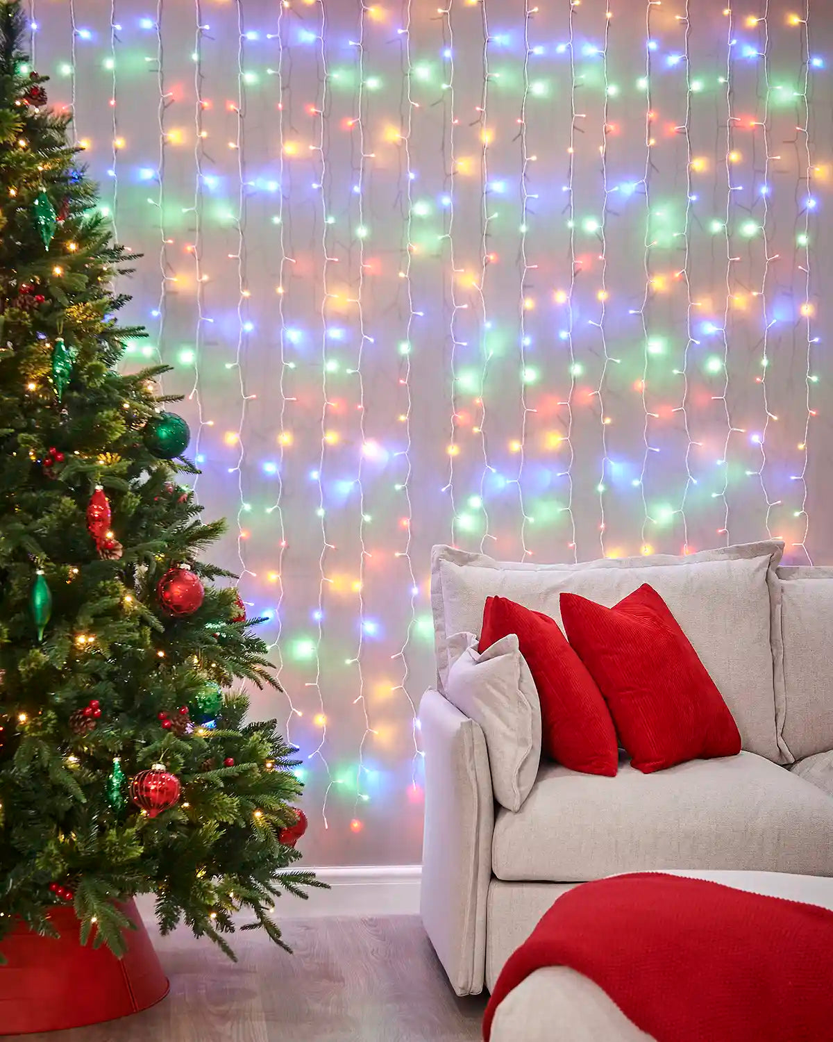 Multi colour curtain lights on white cable displayed on a wall behind a sofa and Christmas tree.