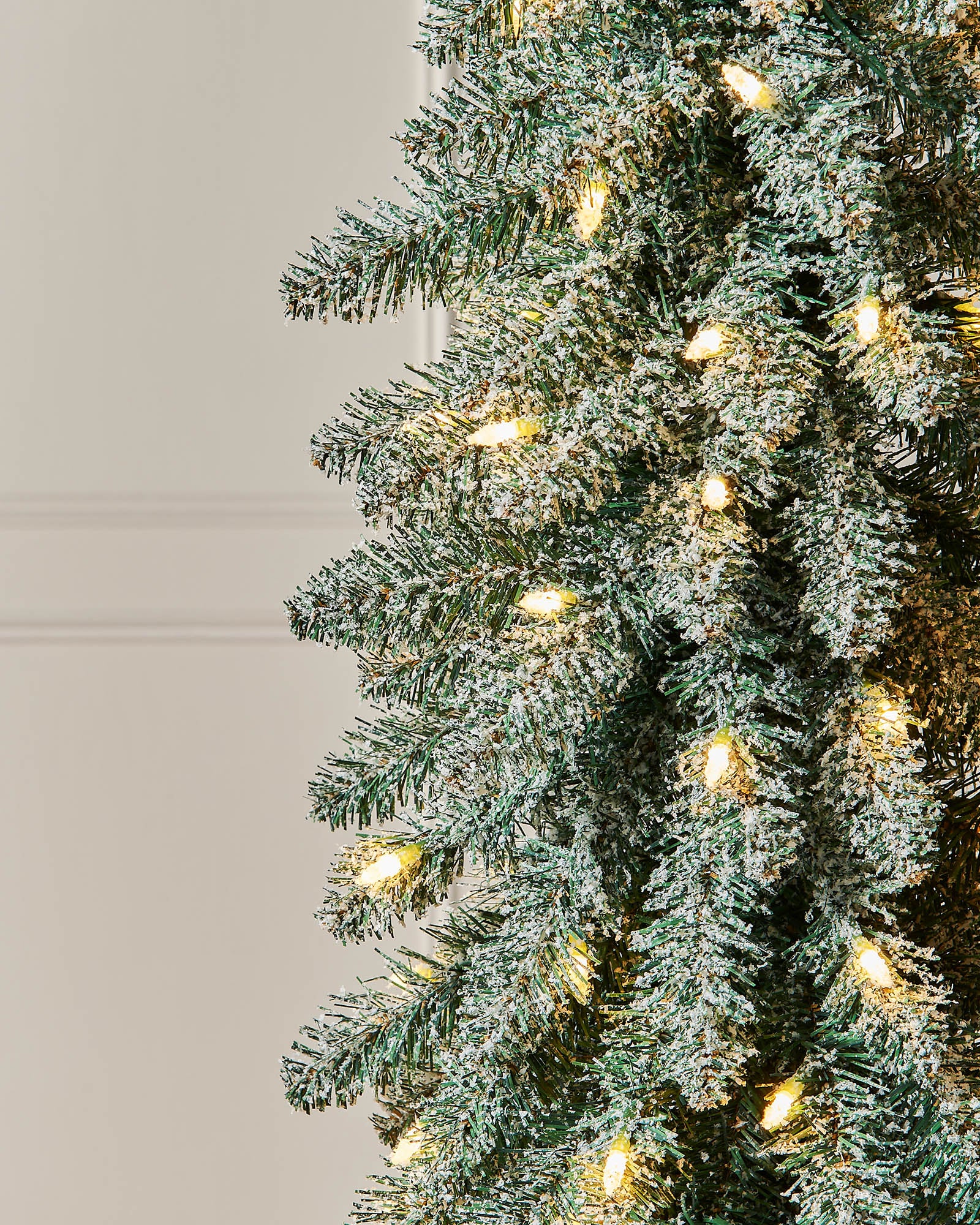 Tall artificial Christmas tree with lights against a white paneled wall.