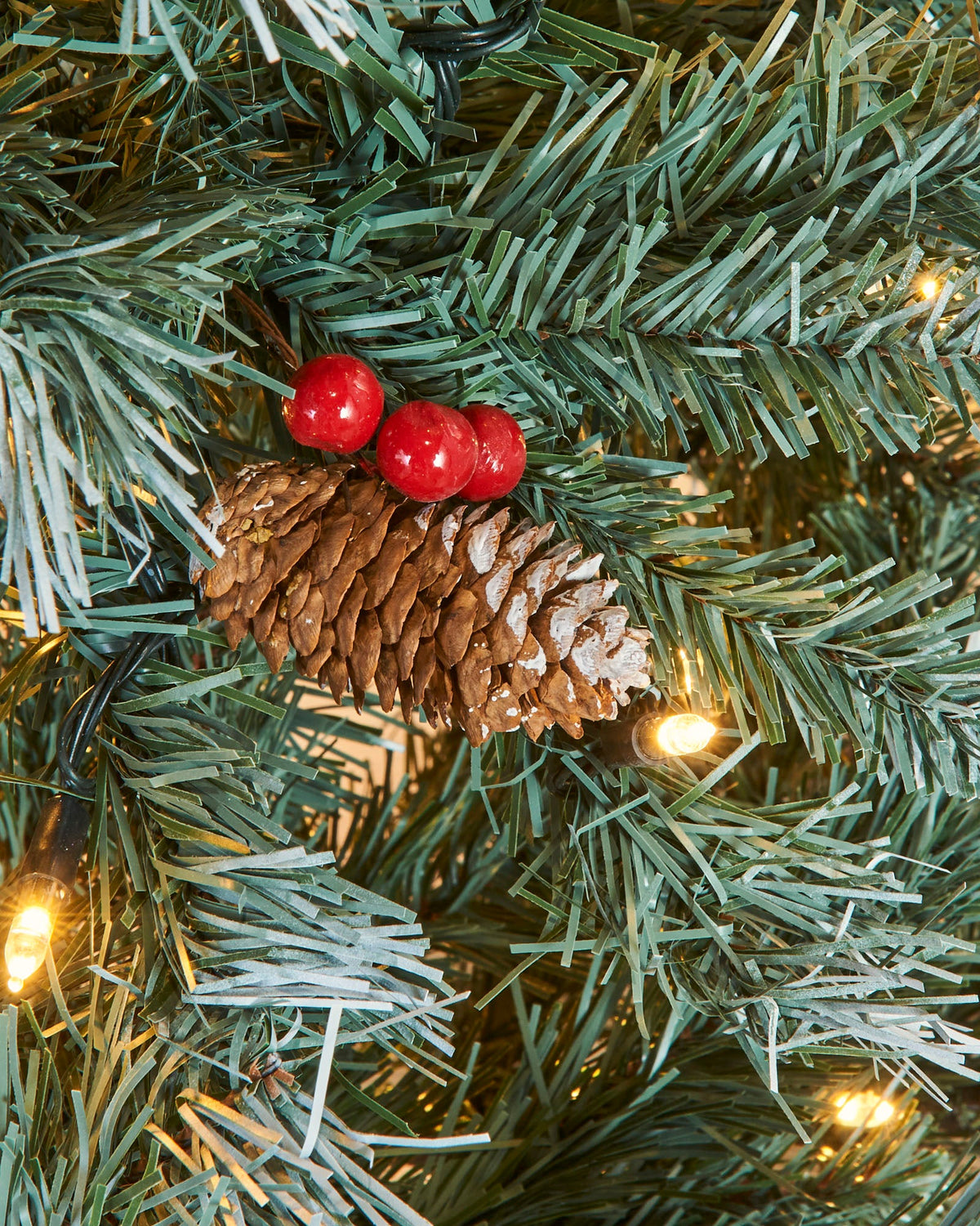 A pre-lit Scandinavian spruce Christmas tree with frosted green PVC branches, warm white LED lights, natural pinecones, and red berries. Positioned in a festive indoor setting with Christmas presents and a seasonal sign, capturing the rustic charm of a snow-dusted Nordic forest.