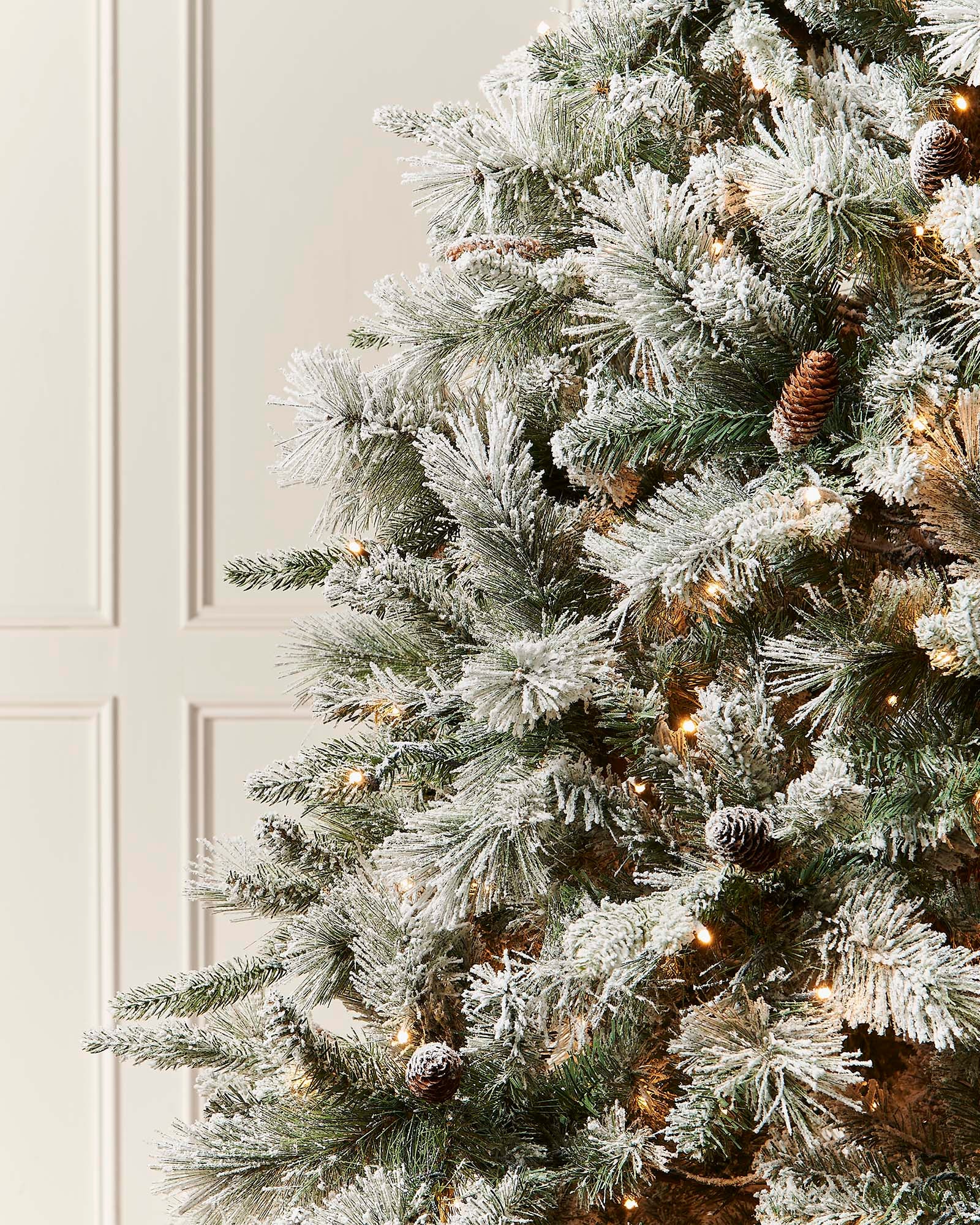 Frosted Christmas tree with pinecones and lights against a neutral background.