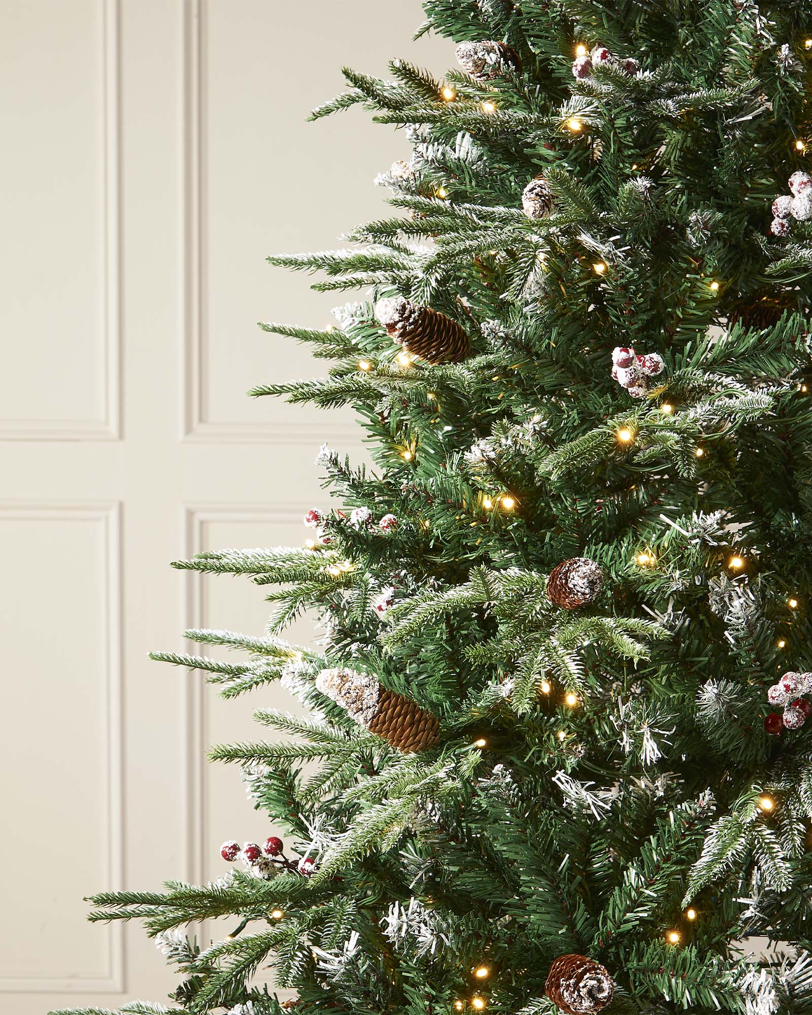 Pre-lit frosted Norwegian Spruce Christmas tree with snow-dusted branches, natural pinecones, and red berries. Features warm white multi-function LED lights and a lifelike PE and PVC tip construction.