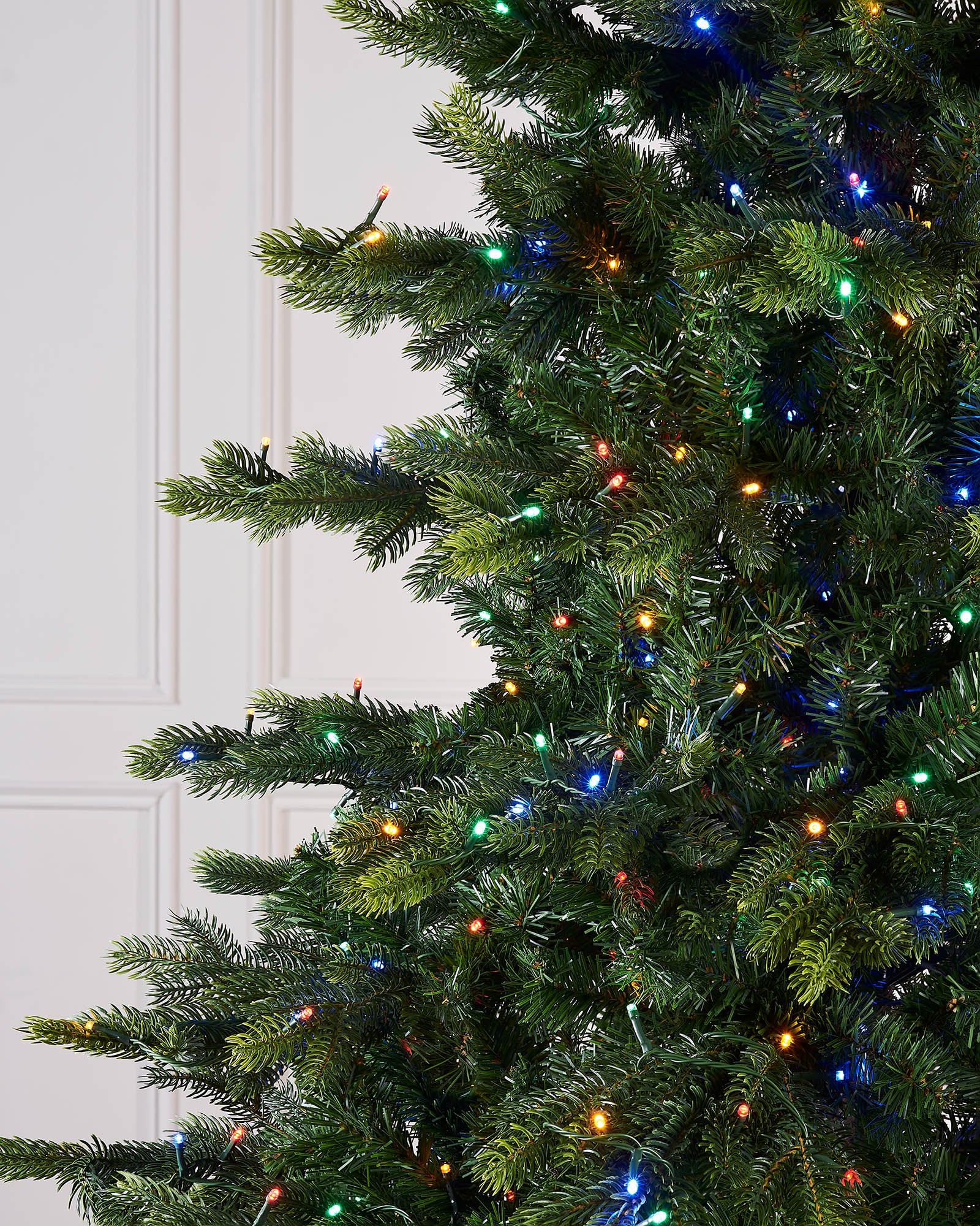 Close-up of a Christmas tree with colorful lights against a white paneled background.