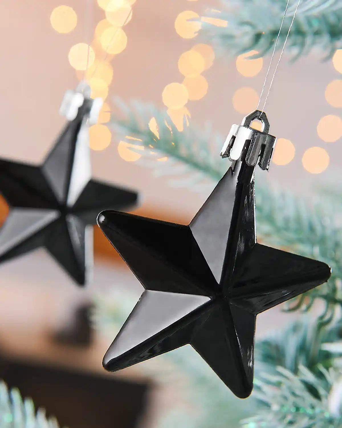 2 gloss Black star hanging baubles on a snow flocked Christmas tree with blurry lights in background 