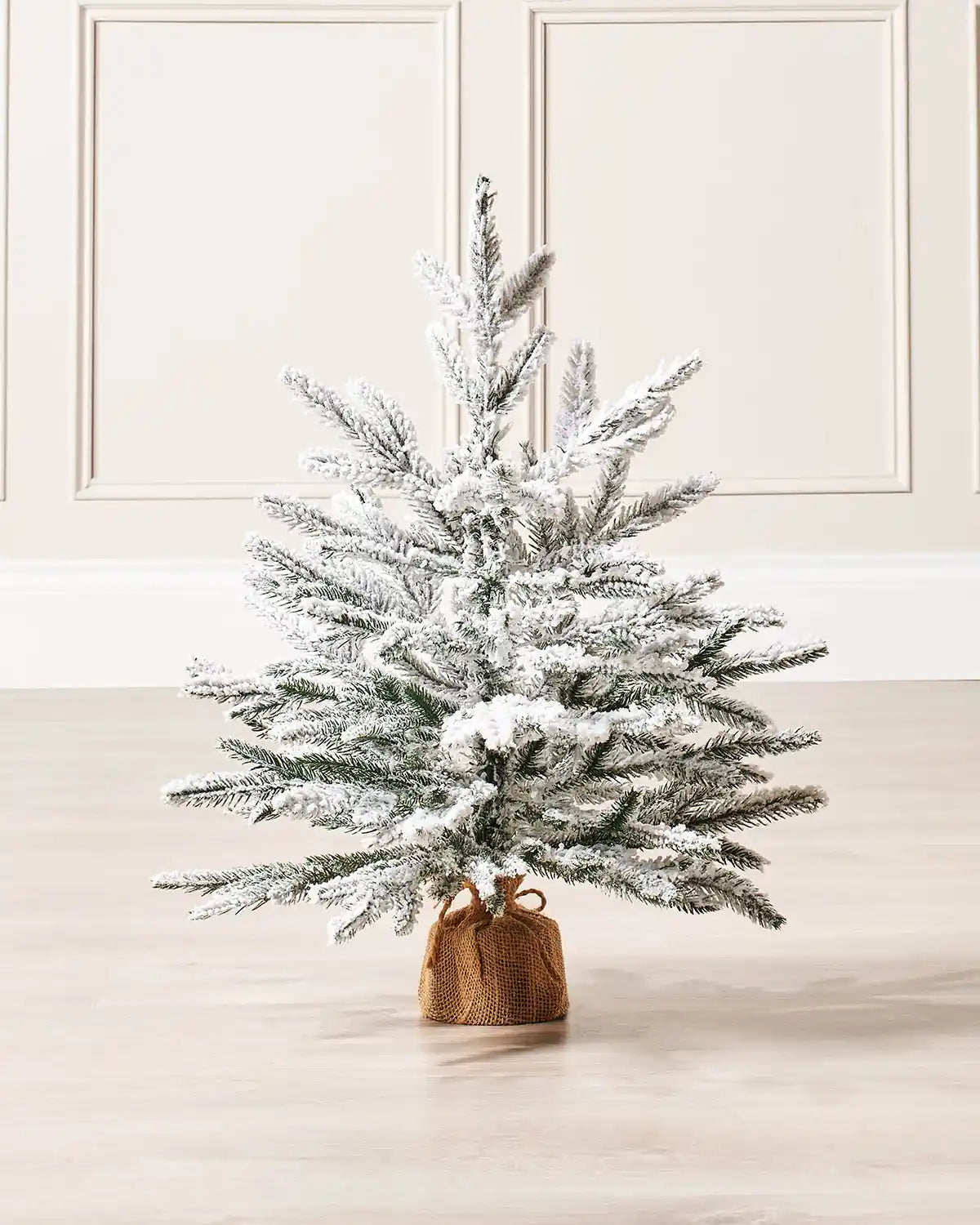 2ft Snow flocked artificial Christmas tree with a brown hessian-wrapped base on a cream panel textured background