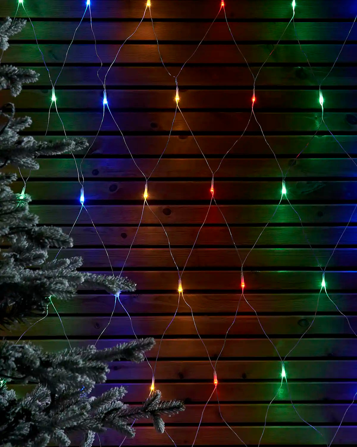 Multi-colour net lights displayed in a diamond pattern against a wooden wall beside a frosted tree.