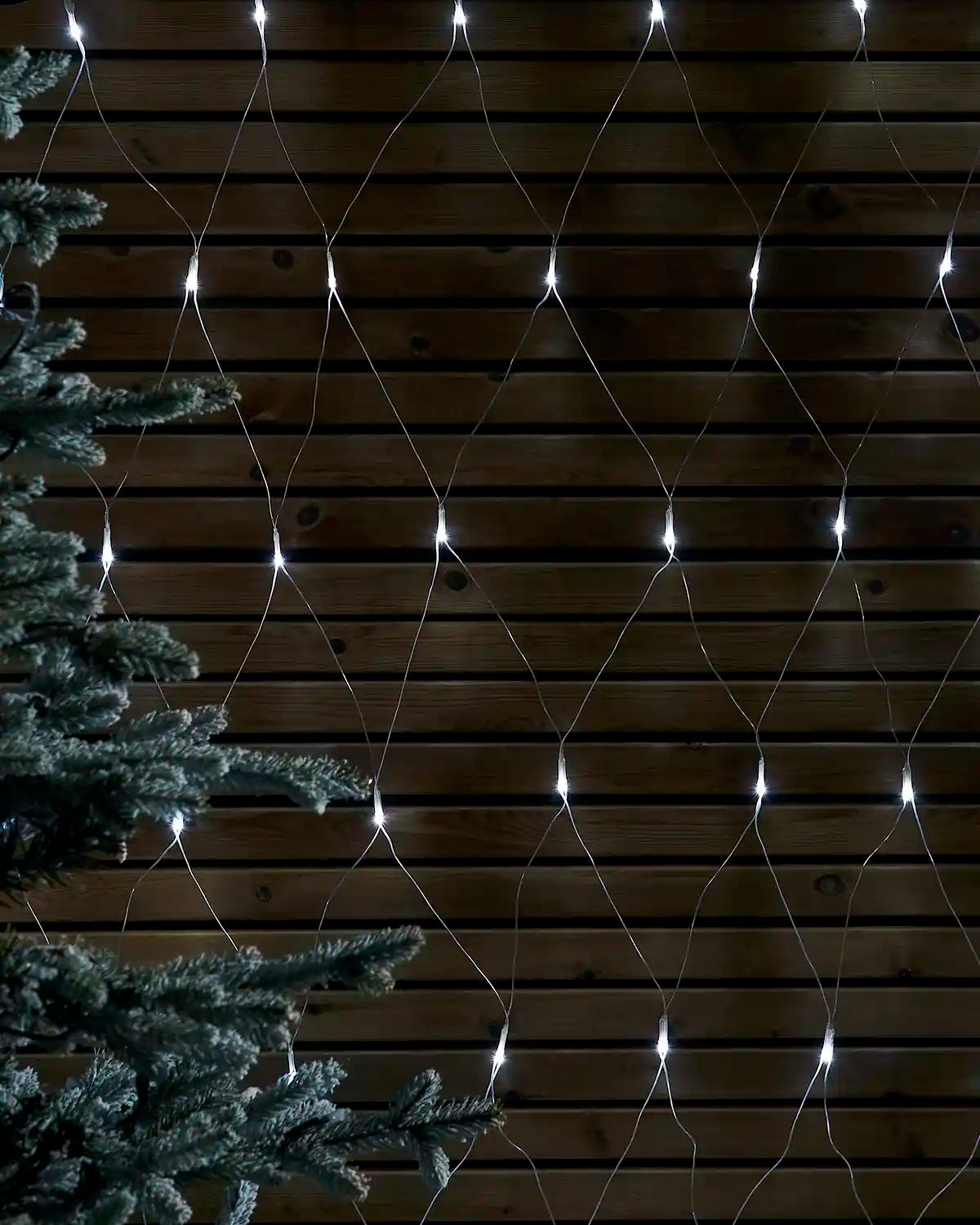 White net lights displayed in a diamond pattern against a wooden wall beside a frosted tree.