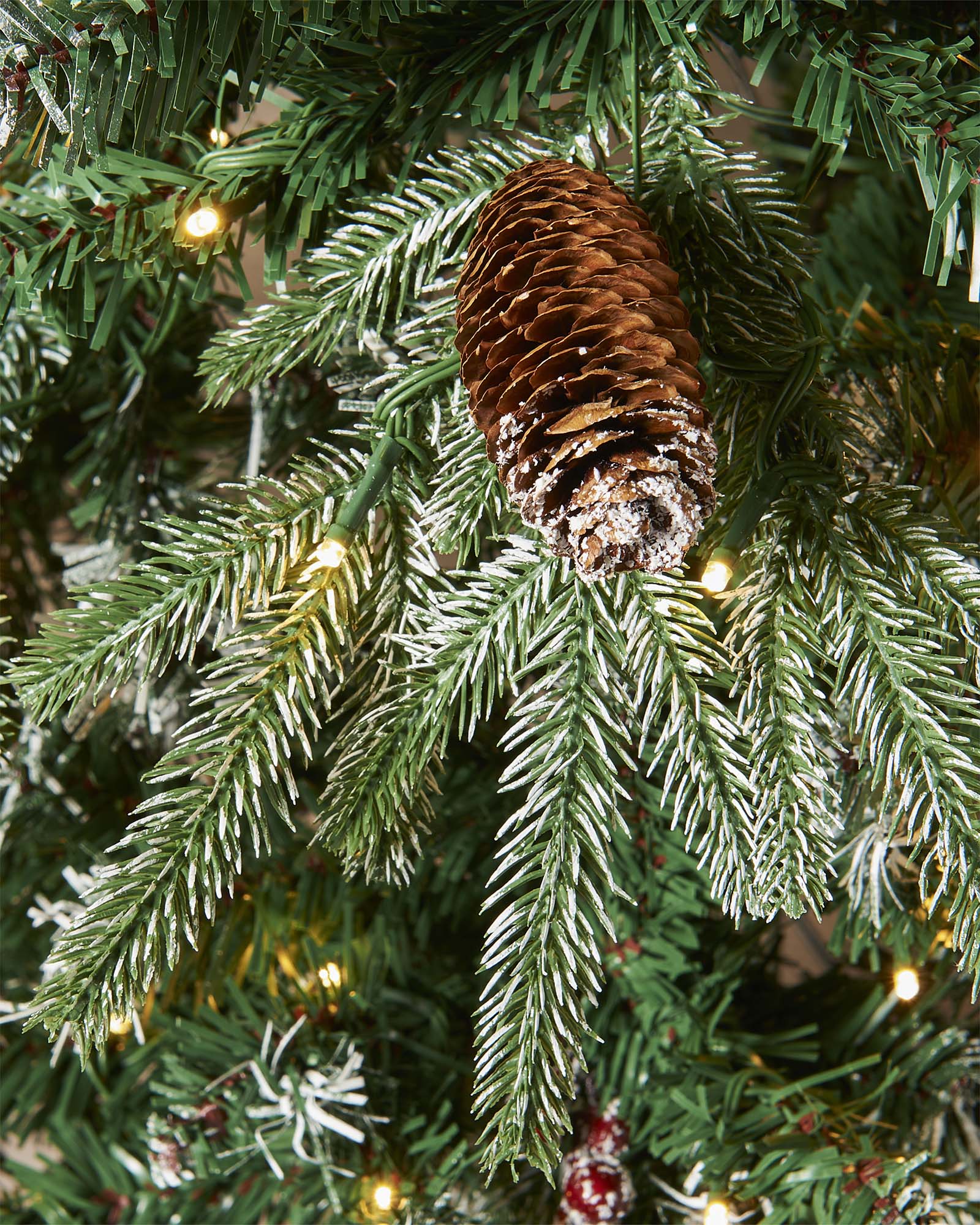 Pre-lit frosted Norwegian Spruce Christmas tree with snow-dusted branches, natural pinecones, and red berries. Features warm white multi-function LED lights and a lifelike PE and PVC tip construction.