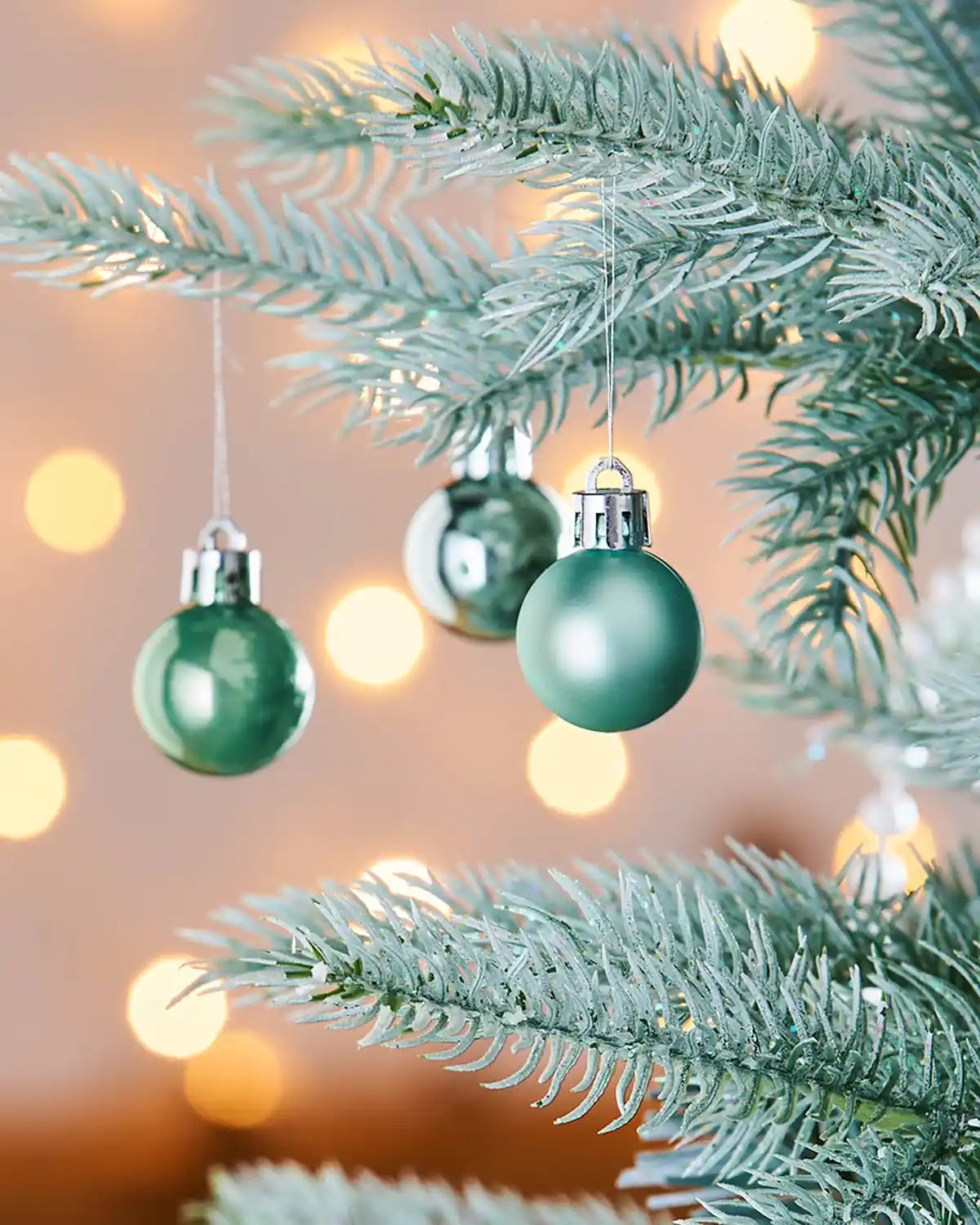 3 Green Christmas baubles hanging on a green tree branch with blurred lights in the background