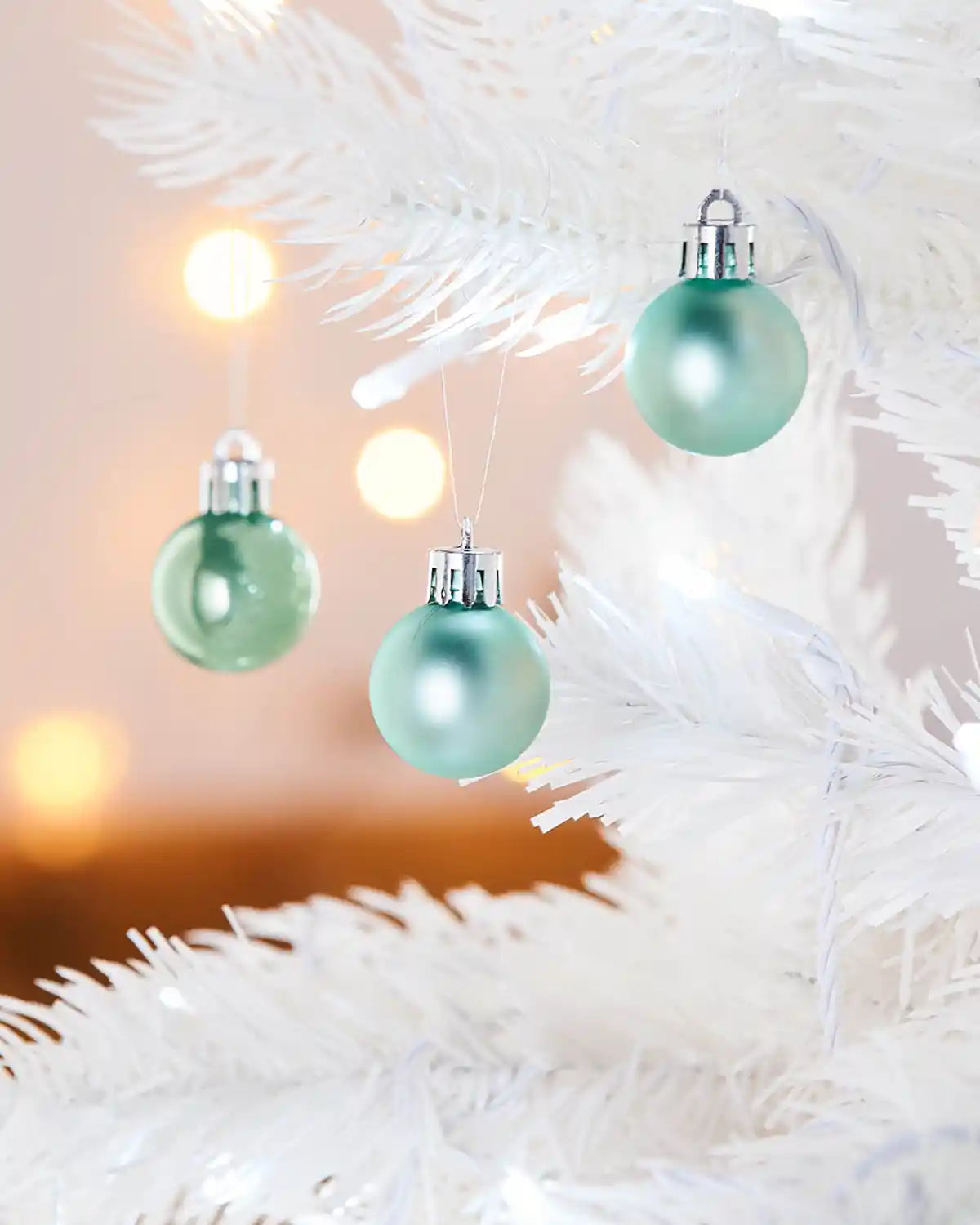 3 Green Christmas baubles hanging on a white tree branch with blurred lights in the background